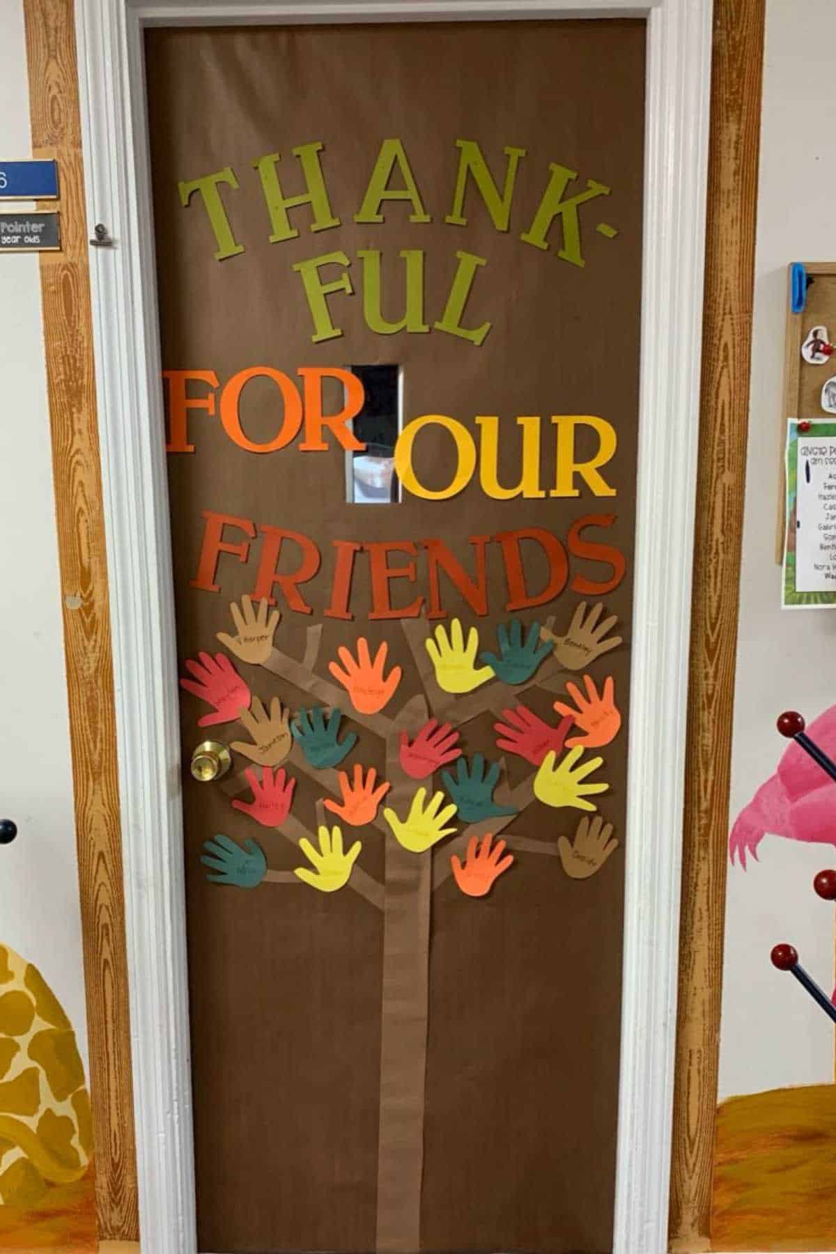 A classroom door decorated with the words "Thankful for our friends" and colorful paper handprints arranged as leaves on a tree.