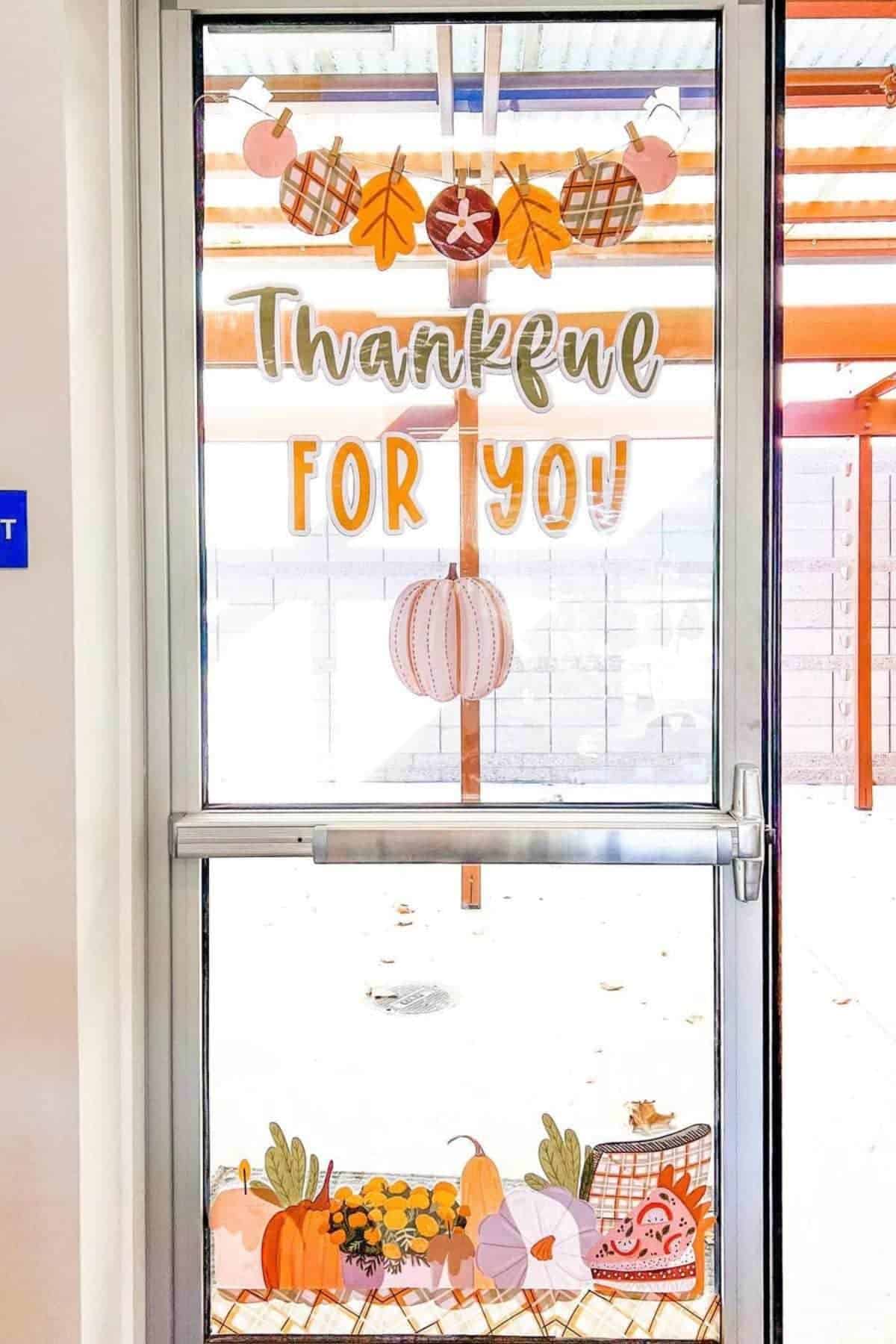 Glass door decorated with autumn-themed cutouts, including pumpkins, leaves, and a banner reading “Thankful for You.”.