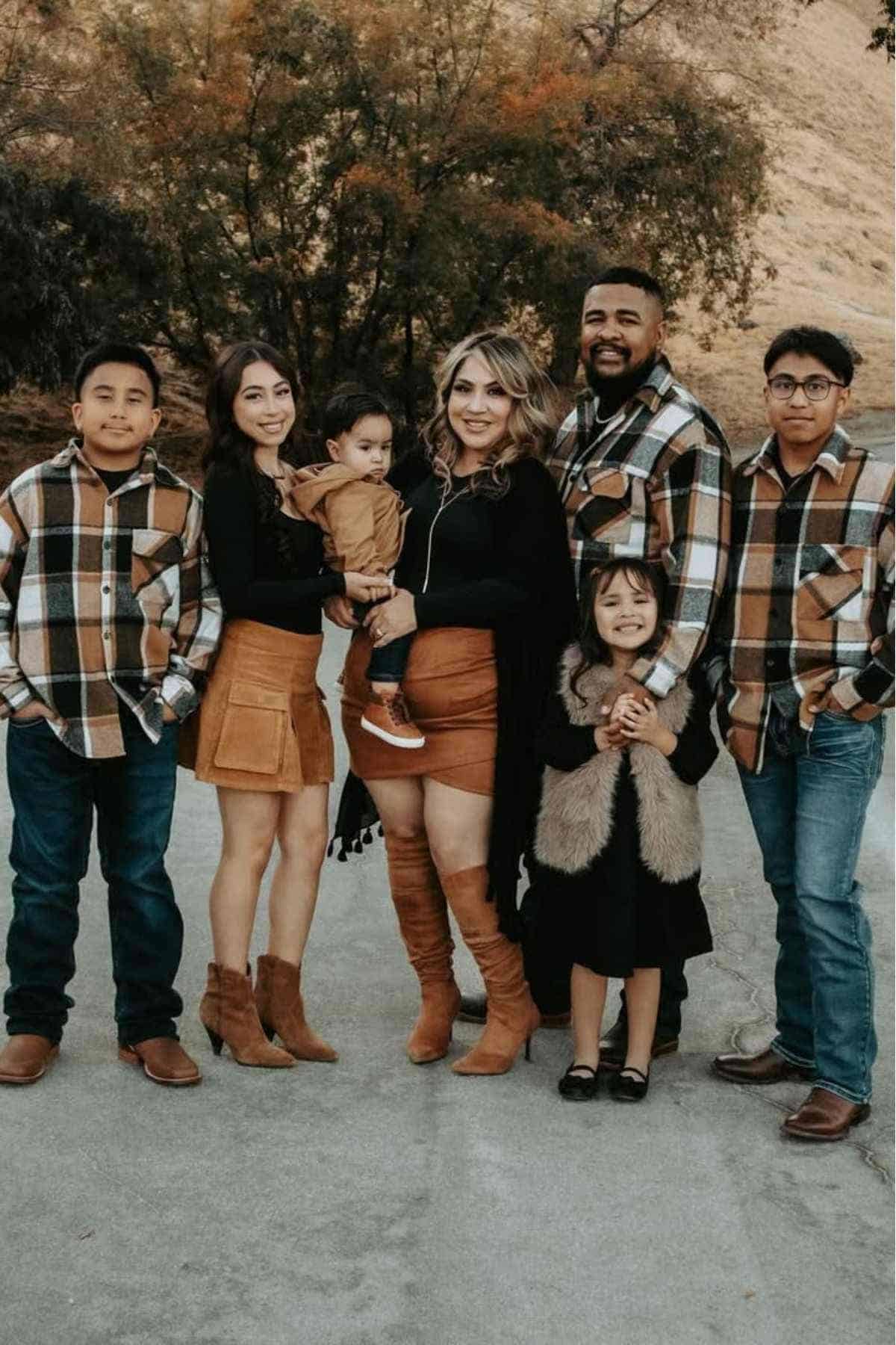 A family of seven poses outdoors, wearing coordinated brown, black, and tan outfits. The background shows trees and a hillside.