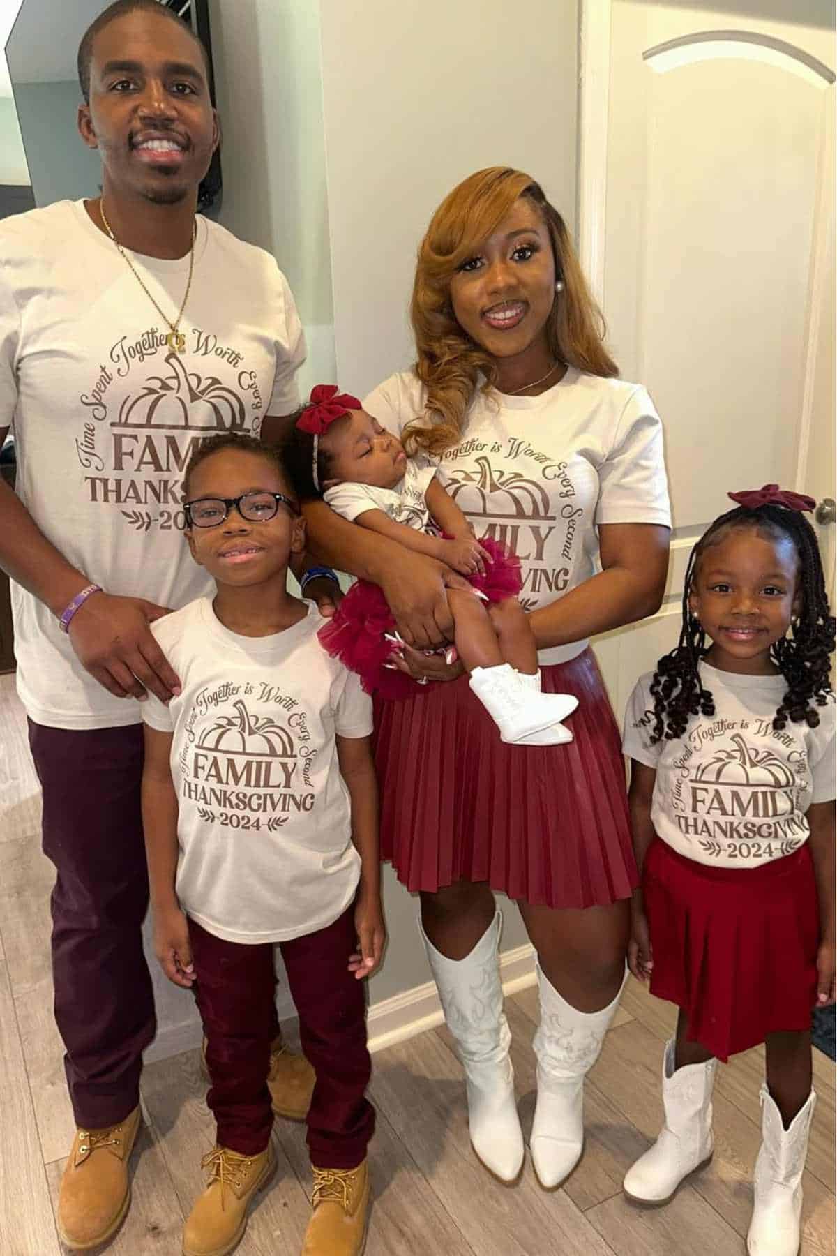 A family of five poses indoors, all wearing matching "Family Thanksgiving 2024" shirts. The parents stand behind their three children, including a baby held by the mother.