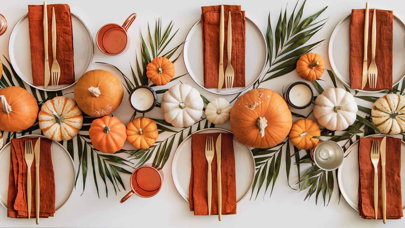 A table set for a meal with orange napkins, gold cutlery, pumpkins, greenery, orange mugs, and candles arranged as a centerpiece.