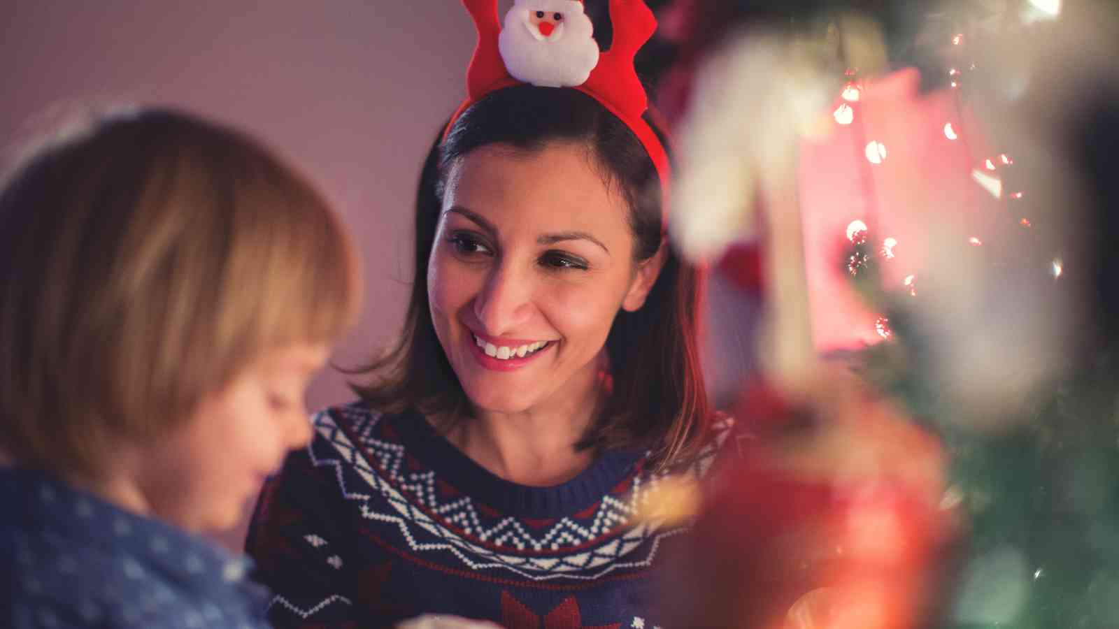 A woman wearing reindeer antlers with a Santa figure smiles at a child near a decorated Christmas tree.