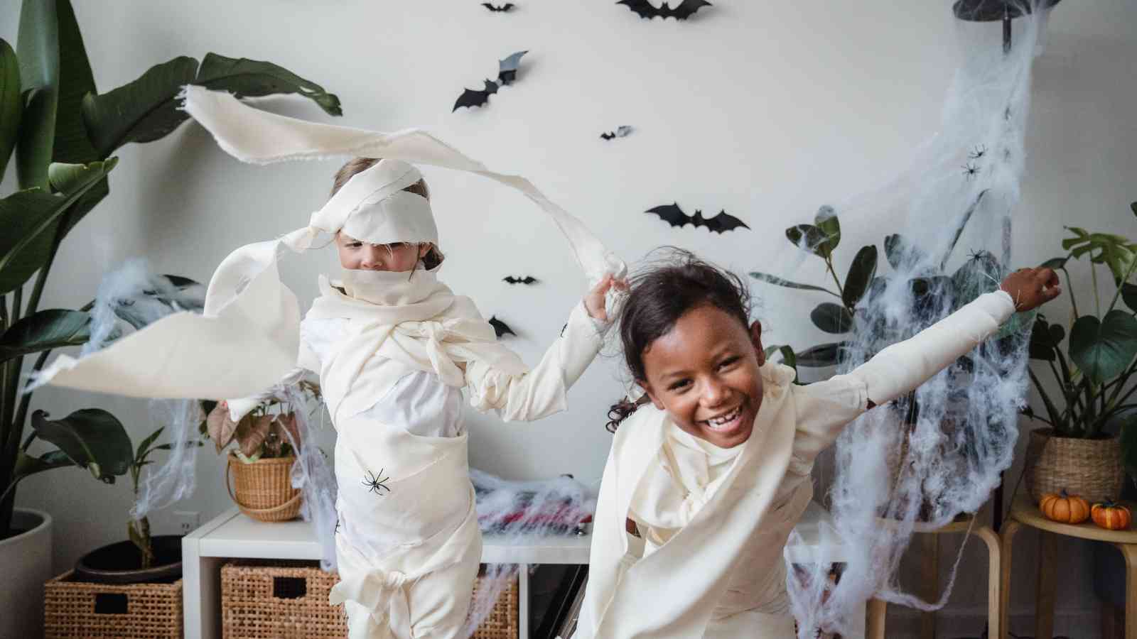 Two children dressed as mummies play in a decorated room with fake cobwebs, paper bats on the wall, and houseplants in the background.