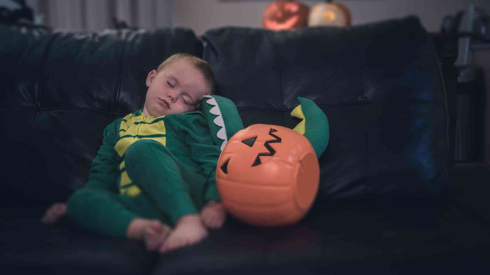 A child in a green dinosaur costume sleeps on a dark sofa next to a pumpkin-shaped Halloween bucket.