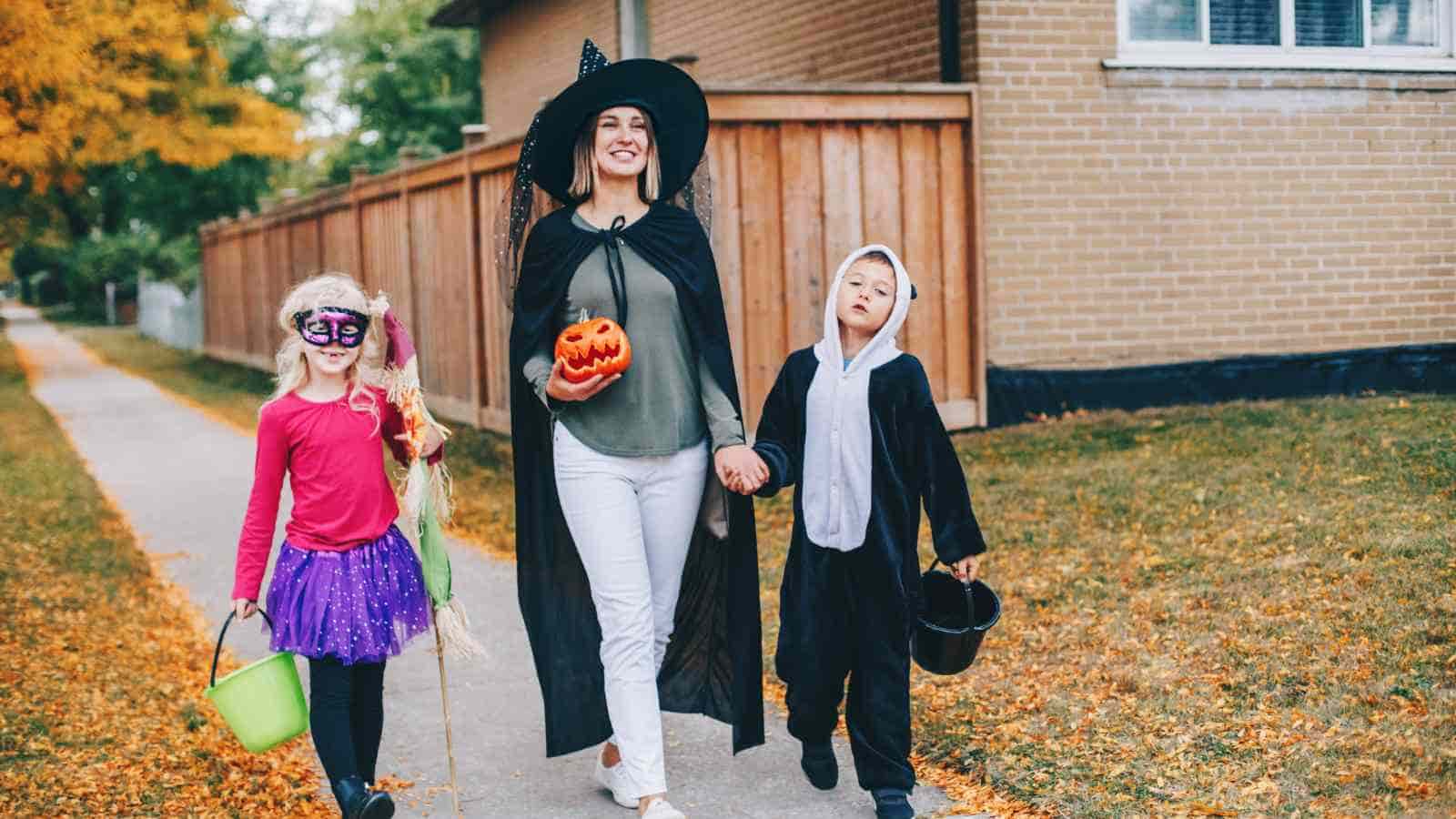 An adult and two children in Halloween costumes walk on a sidewalk. The adult carries a pumpkin bucket; the children hold treat buckets and wear a mask, a tutu, and an animal onesie.