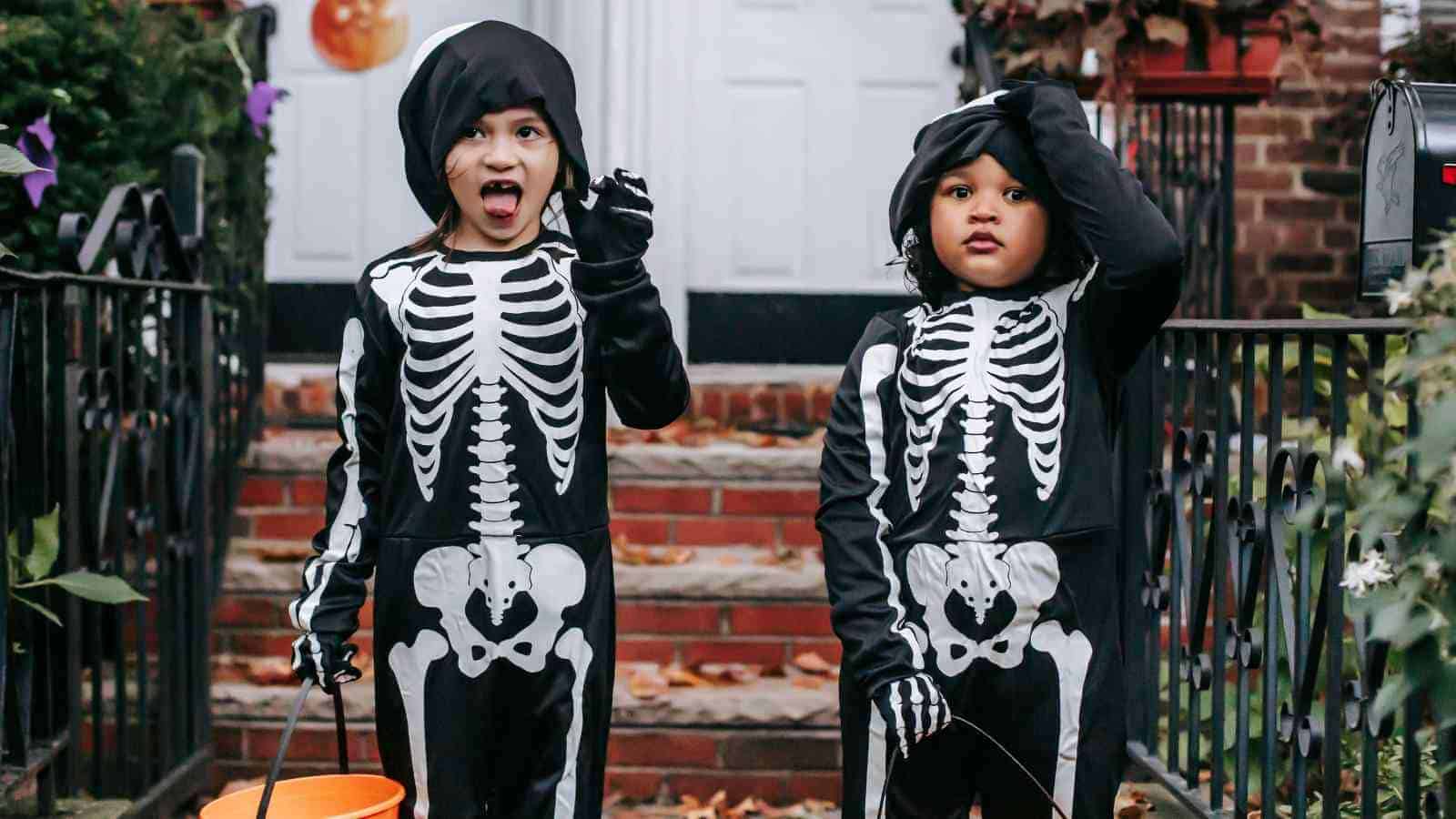 Two children dressed in skeleton costumes stand on a porch, holding orange buckets, likely trick-or-treating for Halloween.