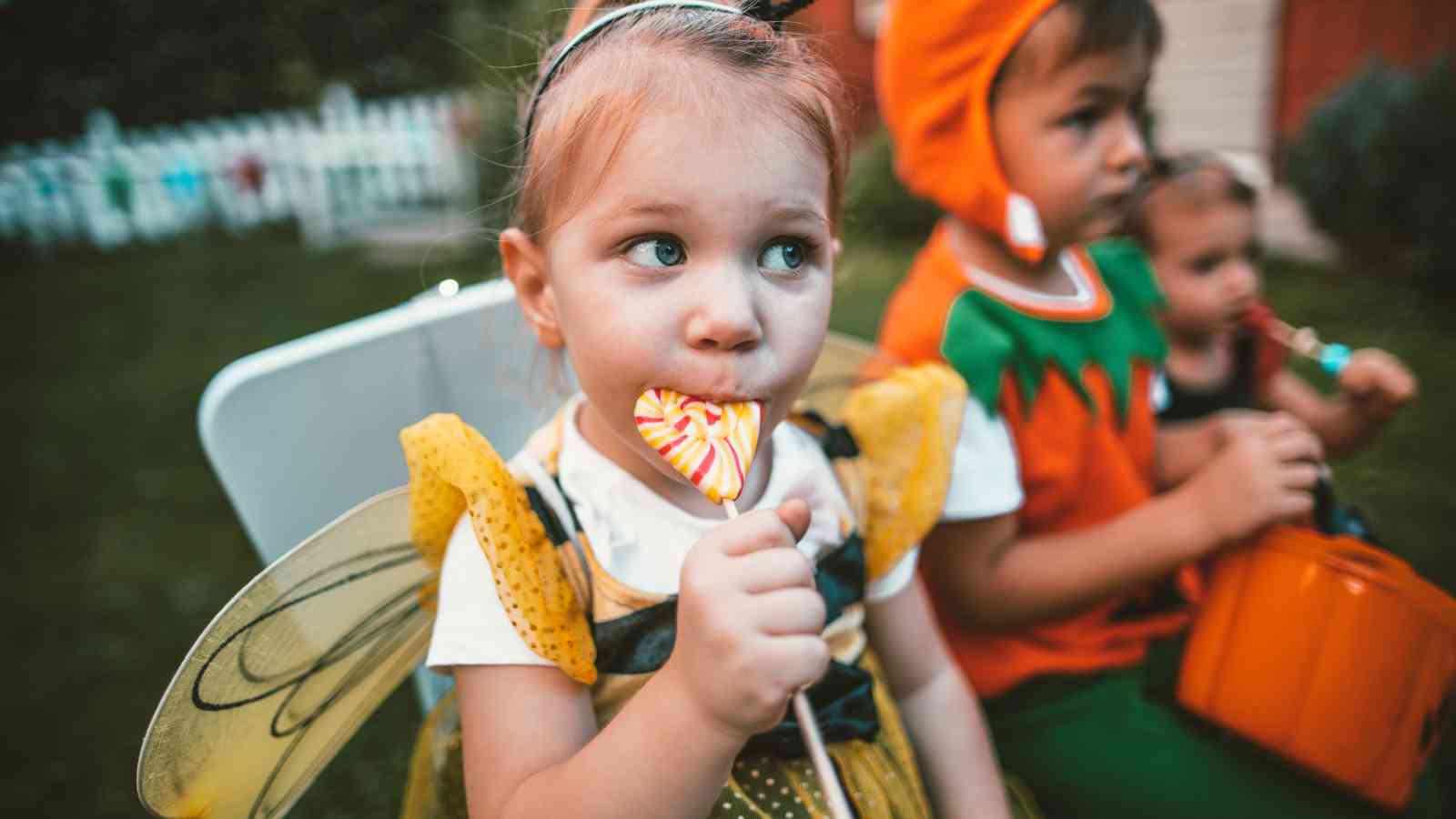 A young child in a bee costume eats a lollipop, sitting next to two other children in costumes, outdoors.