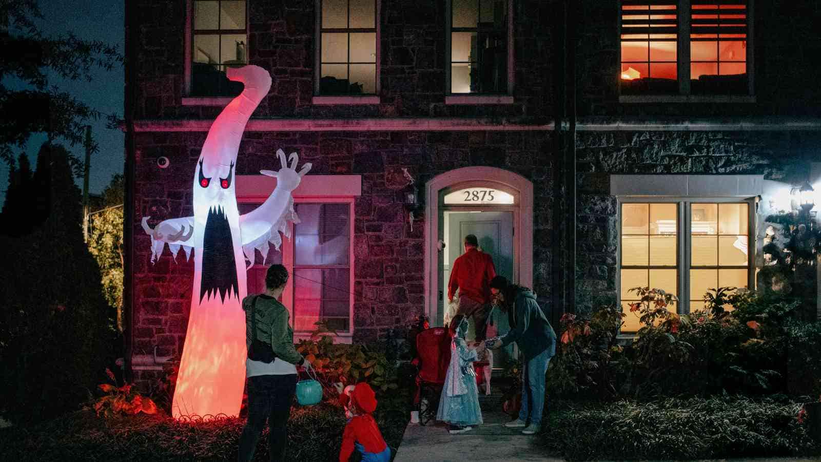 Children in costumes trick-or-treat at a house decorated with a large inflatable ghost and Halloween lights at night.