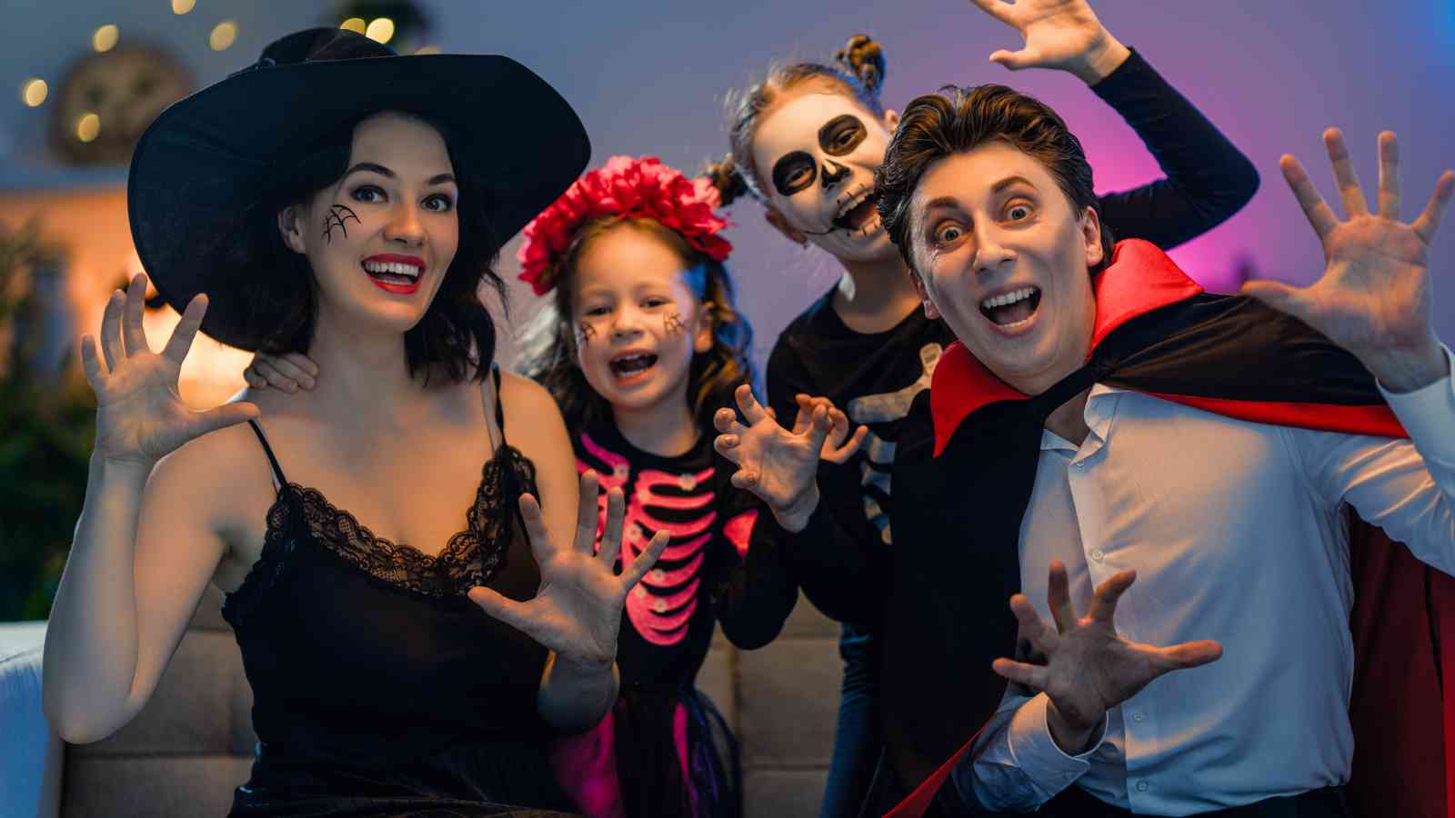 Four people in Halloween costumes, including a witch, a vampire, and two children with face paint and skeleton outfits, pose together making playful scary faces.