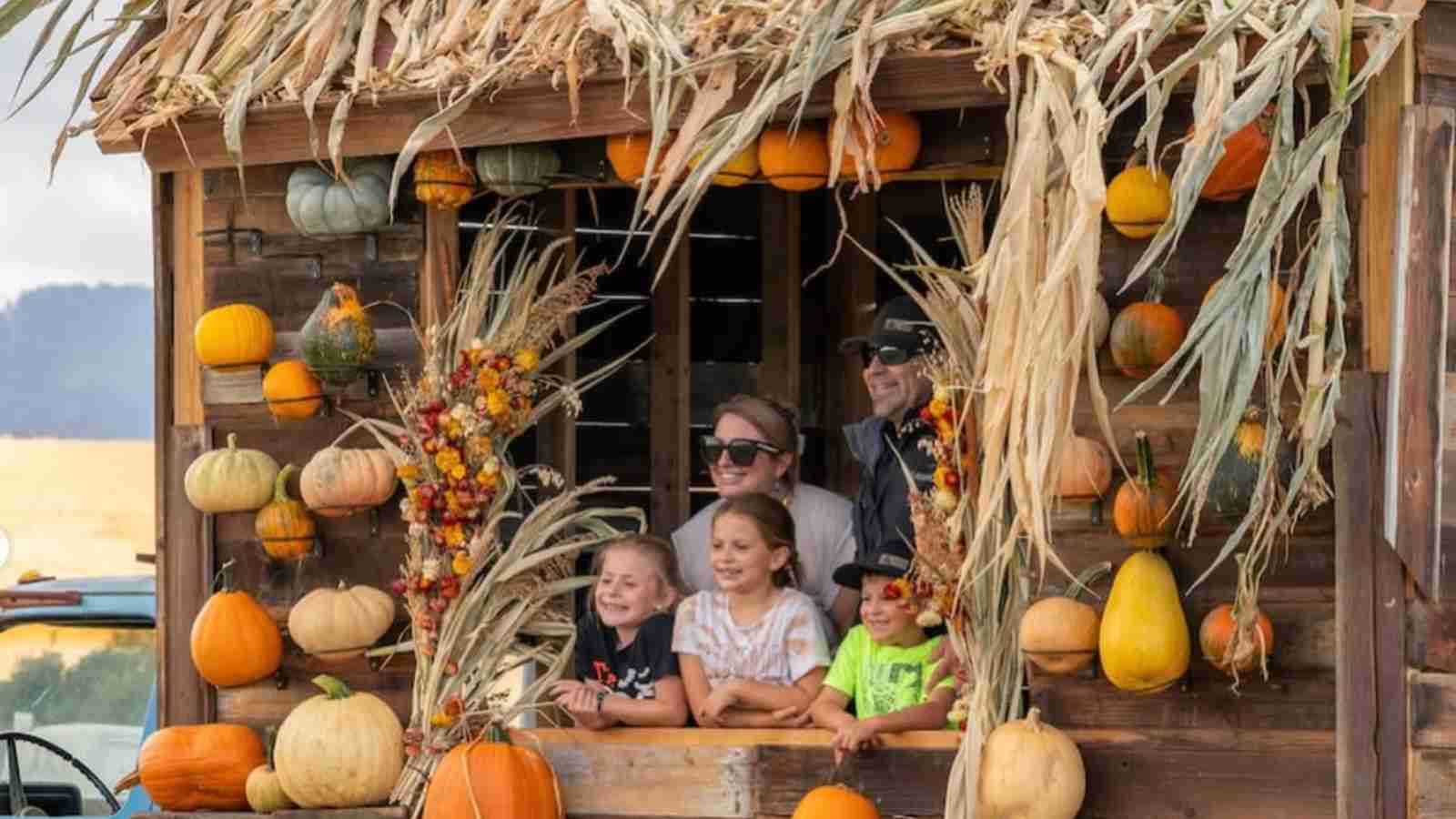 A group of people, including children, smiles from the window of a wooden structure decorated with pumpkins, gourds, and dried corn stalks.