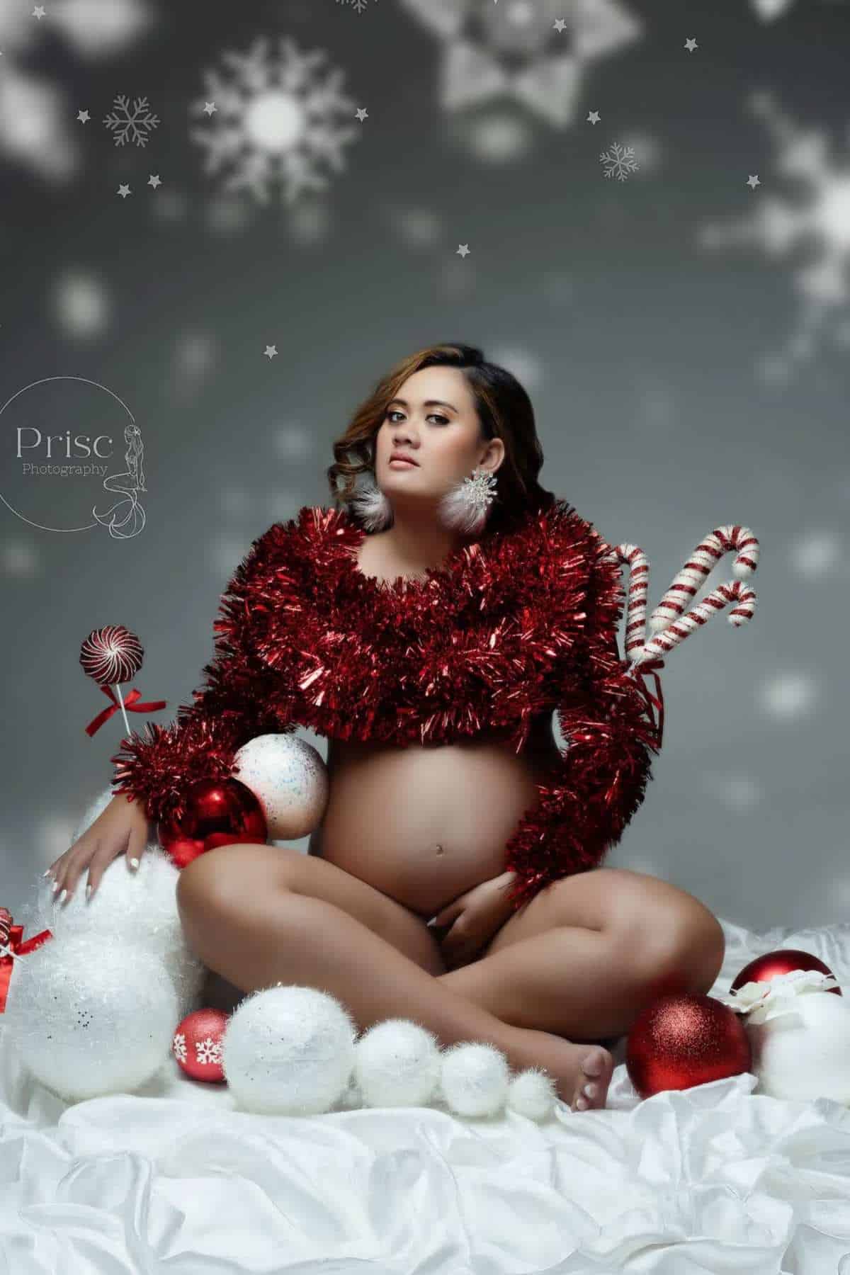 A pregnant woman sits on white fabric, wearing a red tinsel top and festive earrings, surrounded by holiday decorations and candy canes, with snowflake graphics in the background.