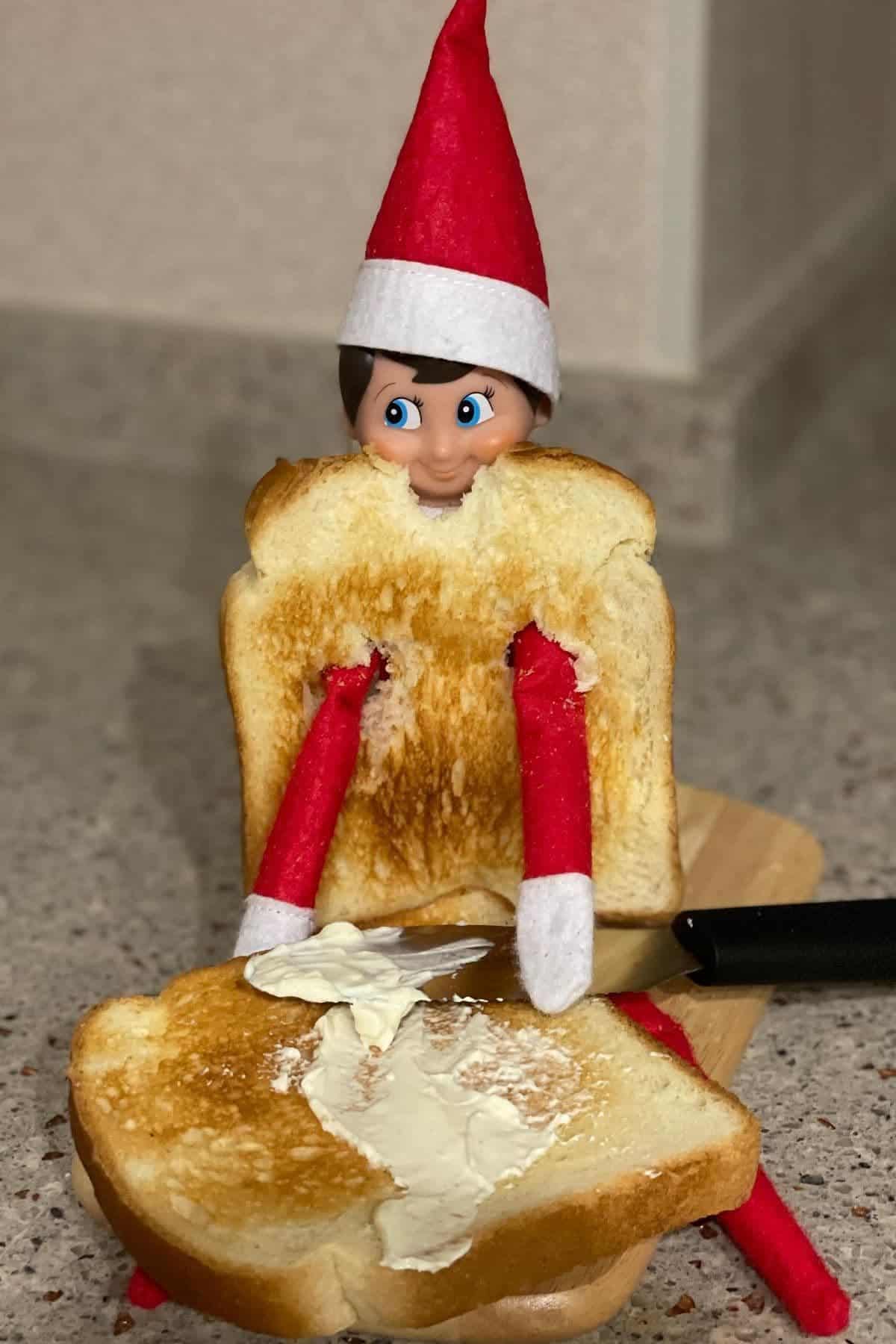 A doll dressed as an elf sits between two slices of toast, with a knife spreading butter on the front slice, on a kitchen counter.