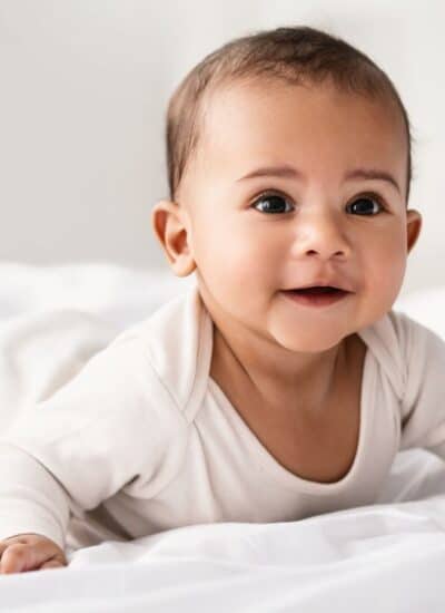 A baby wearing a light-colored onesie lies on its stomach on a white bed, looking forward with a neutral expression.
