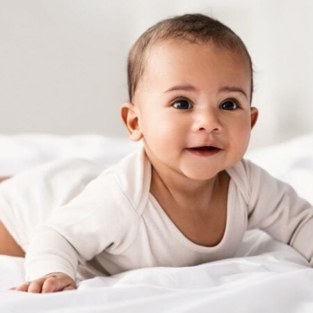 A baby wearing a light-colored onesie lies on its stomach on a white bed, looking forward with a neutral expression.