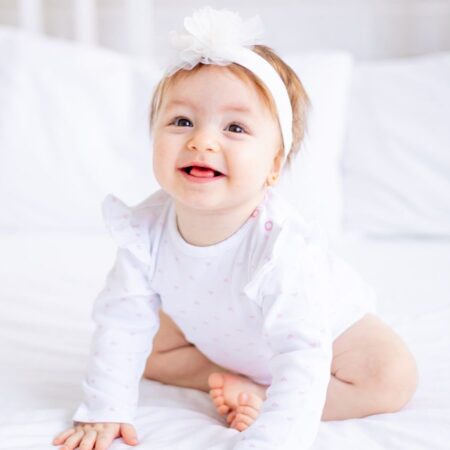 Baby wearing a white headband and white outfit sitting and smiling on a bed with white pillows and bedding.