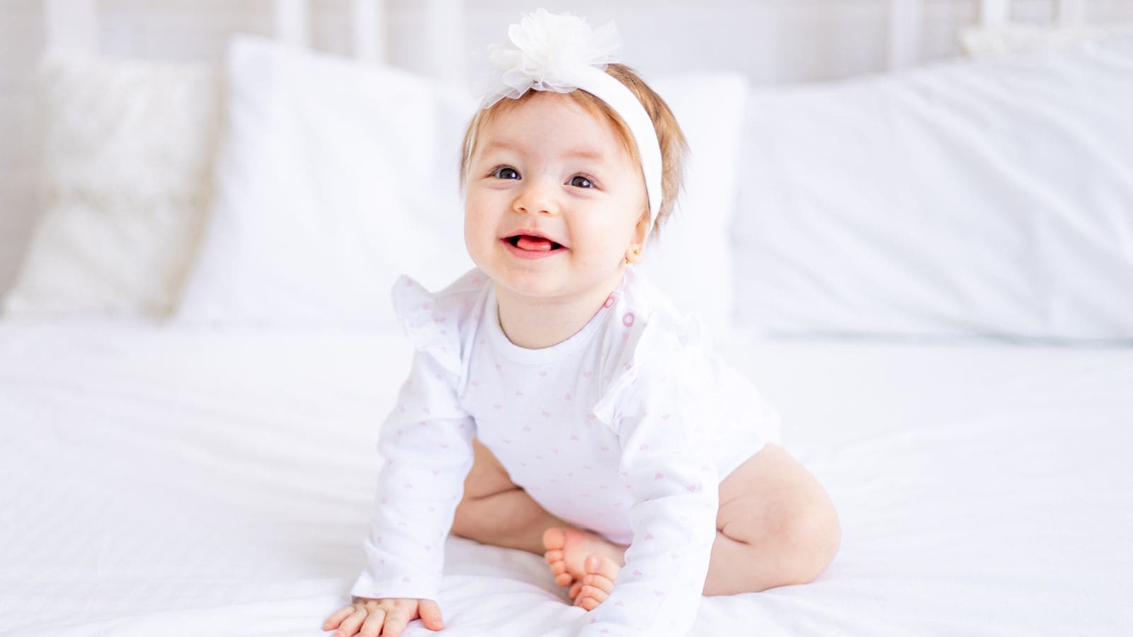 Baby wearing a white headband and white outfit sitting and smiling on a bed with white pillows and bedding.