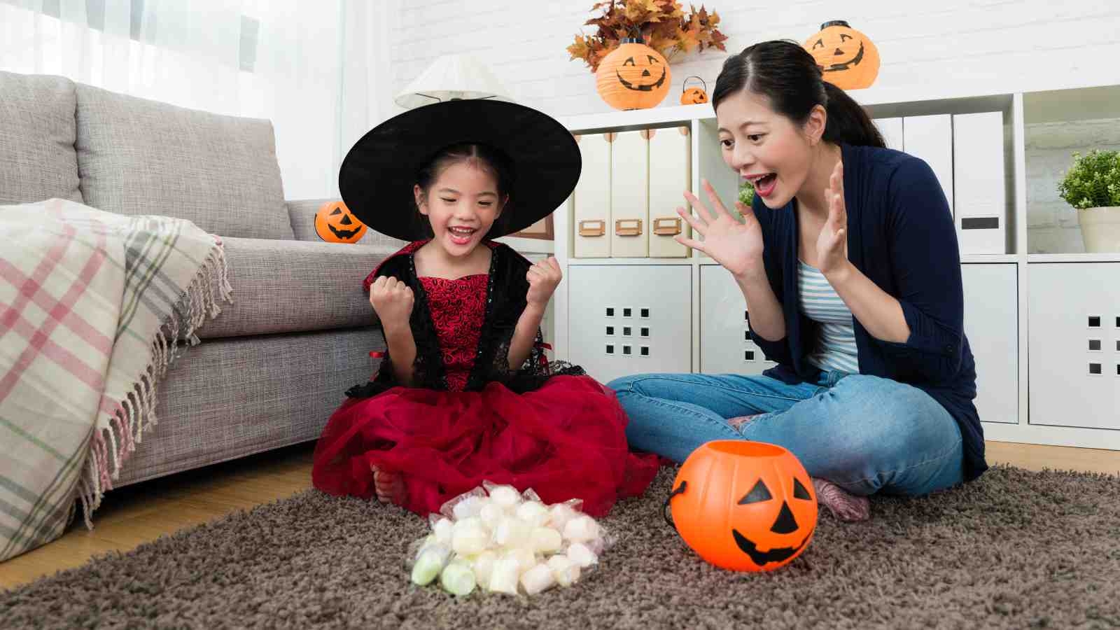 A child in a witch costume and an adult sit on the floor with Halloween decorations and candy, both appearing excited.