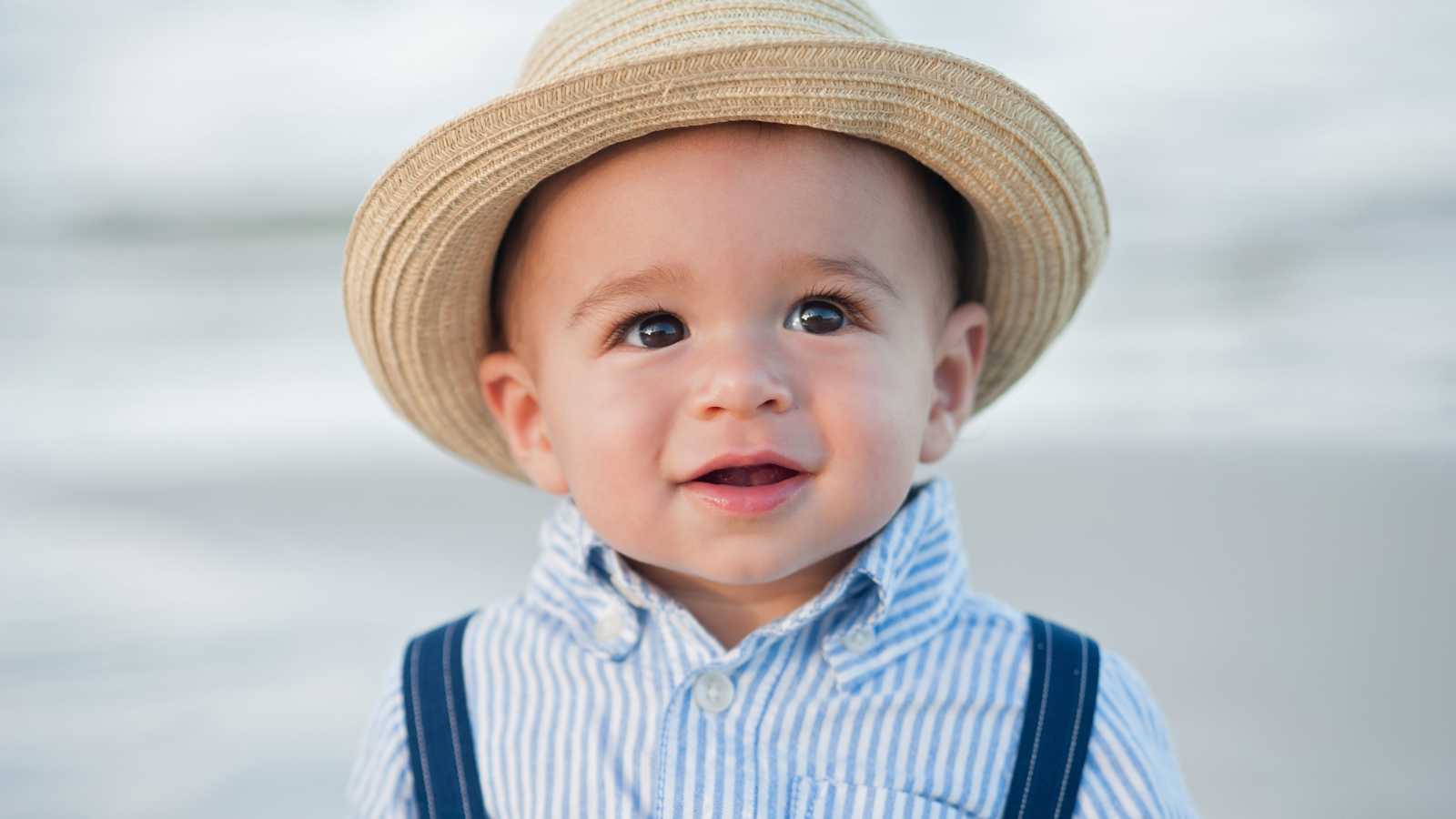 A young child wearing a striped shirt, suspenders, and a straw hat stands outdoors, looking slightly upward with a neutral expression.