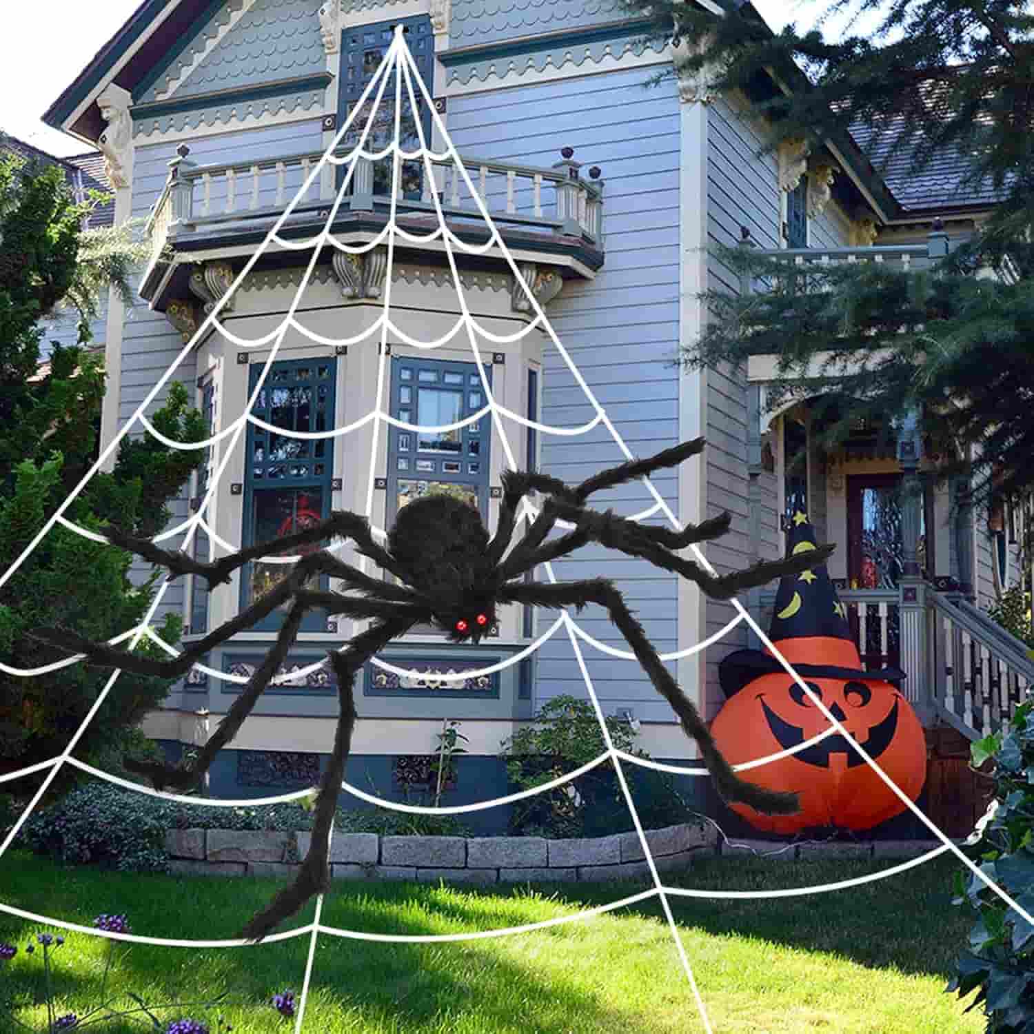 A large fake spider with red eyes sits on a web in front of a house decorated for Halloween, with a pumpkin and witch hat inflatables nearby.