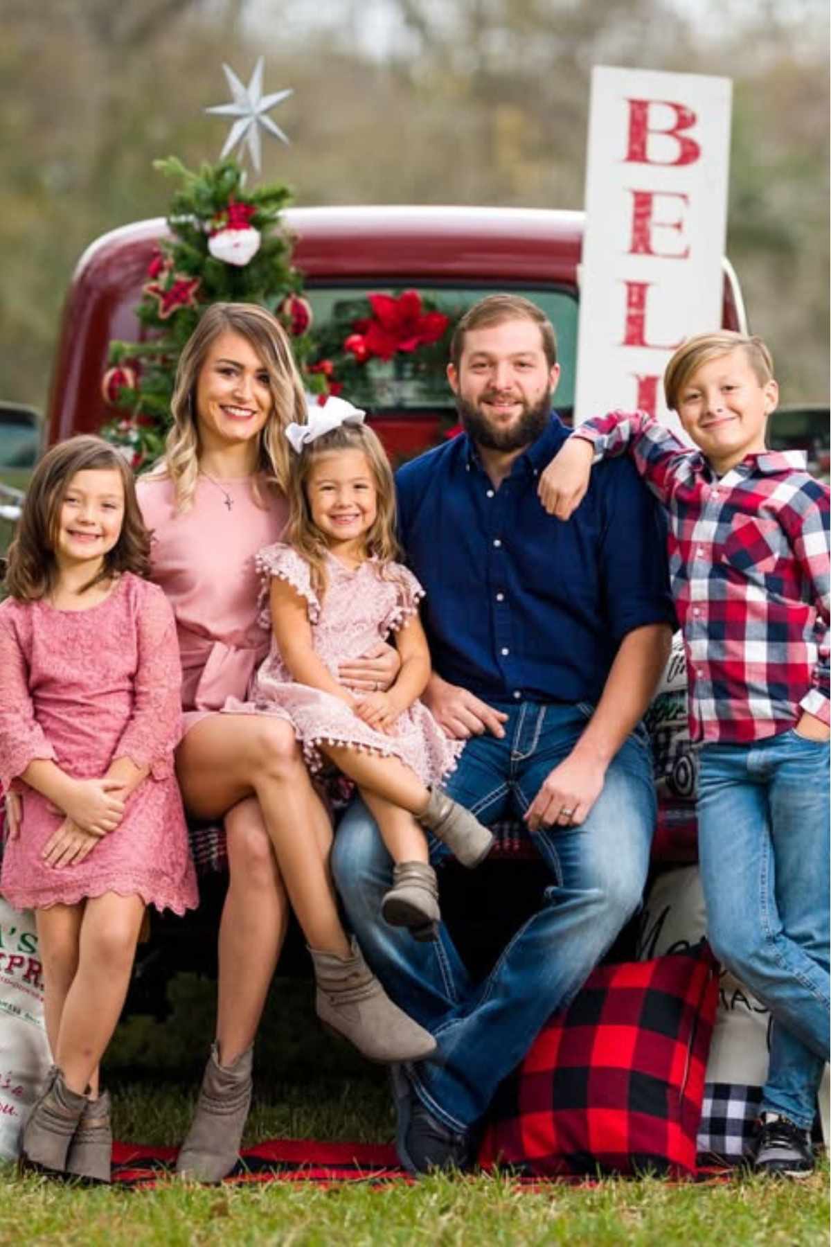 A family of five poses in front of a red truck decorated with a Christmas tree and festive signs, sitting and smiling outdoors.