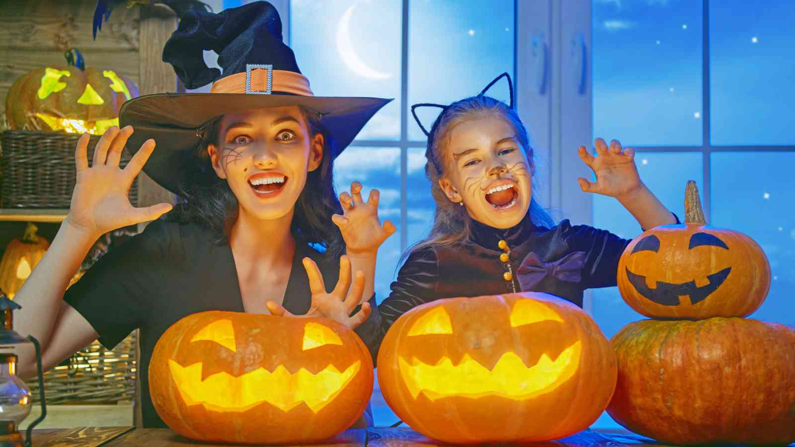 Two people in Halloween costumes pose indoors with carved jack-o’-lanterns on a table, with a night sky showing through the window in the background.