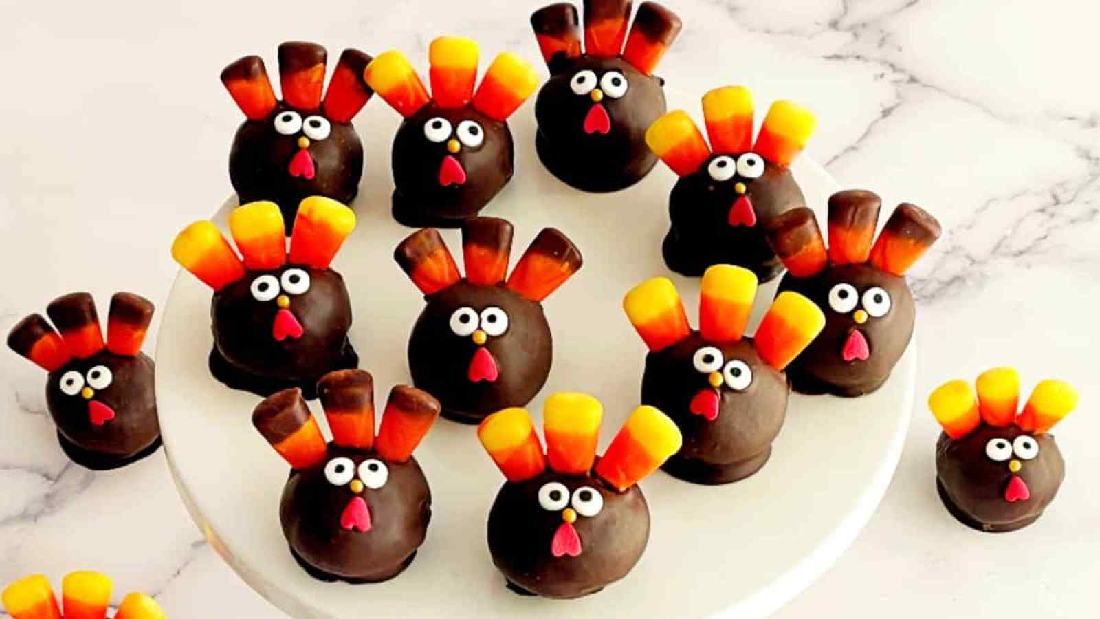 Chocolate-covered cake balls decorated as turkeys with candy corn feathers, candy eyes, and red candy beaks, arranged on a white surface and cake stand.