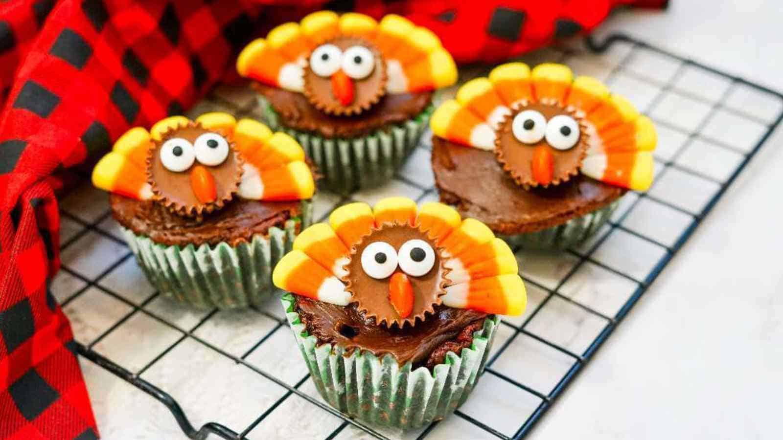 Four chocolate cupcakes decorated with candy to resemble turkeys, featuring candy corn feathers and candy eyes, sit on a cooling rack next to a red and black checkered cloth.
