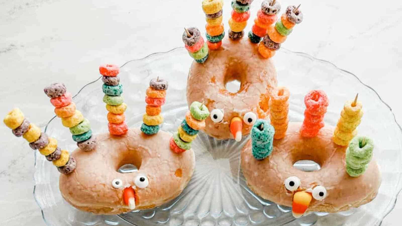 Three donuts decorated as turkeys with candy eyes, candy beaks, and colorful cereal pieces on sticks for feathers, arranged on a clear glass plate.