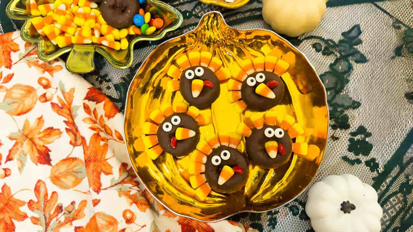 Five turkey-shaped cookies decorated with candy corn and candy eyes are arranged on a gold pumpkin-shaped plate, surrounded by autumn-themed decorations.