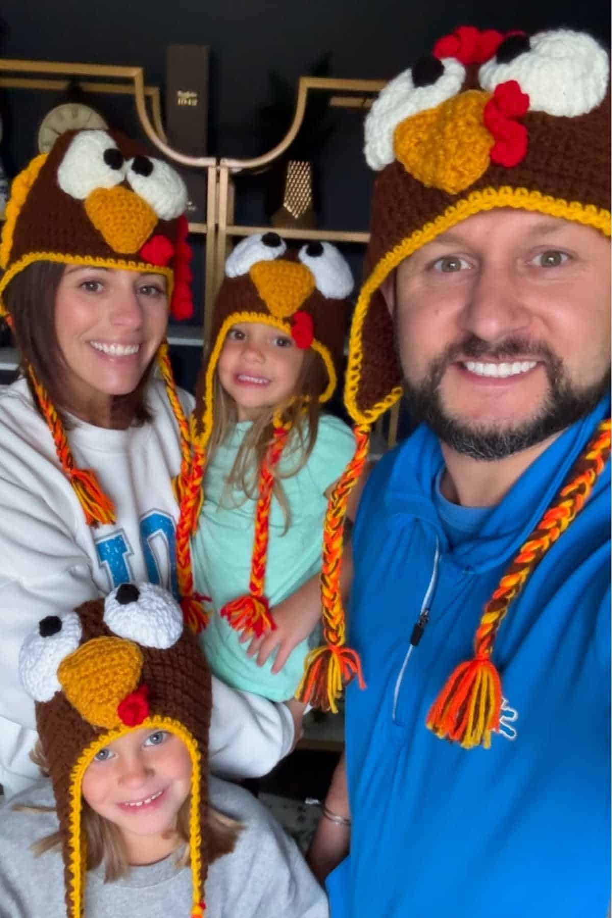 A group of four people, two adults and two children, wearing matching crocheted turkey hats and smiling at the camera.