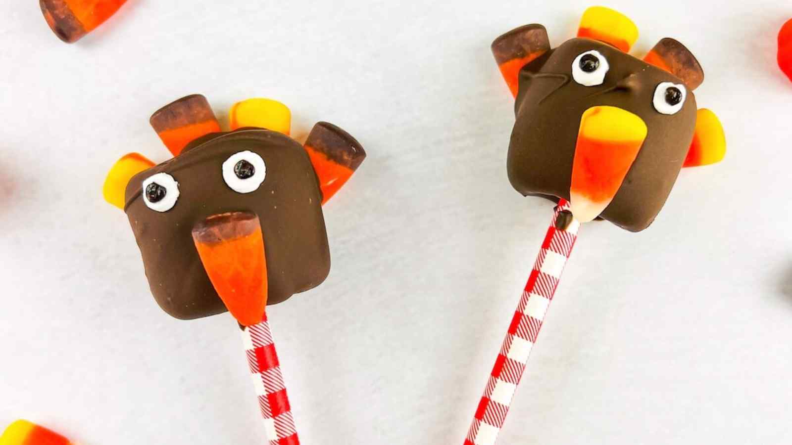 Two marshmallows coated in chocolate, decorated with candy corn and candy eyes to resemble turkeys, on red and white striped sticks.