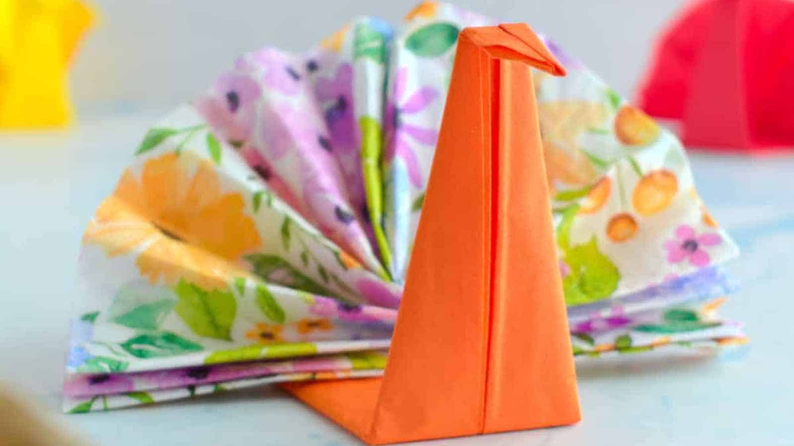 A paper napkin folded into the shape of a peacock, with a colorful floral pattern for the tail and an orange napkin for the body and head.
