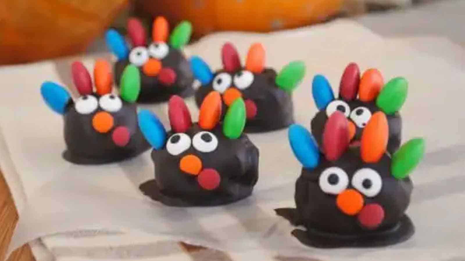 Six small, round chocolate treats decorated with colorful candy pieces to resemble turkeys are arranged on a napkin-covered surface.
