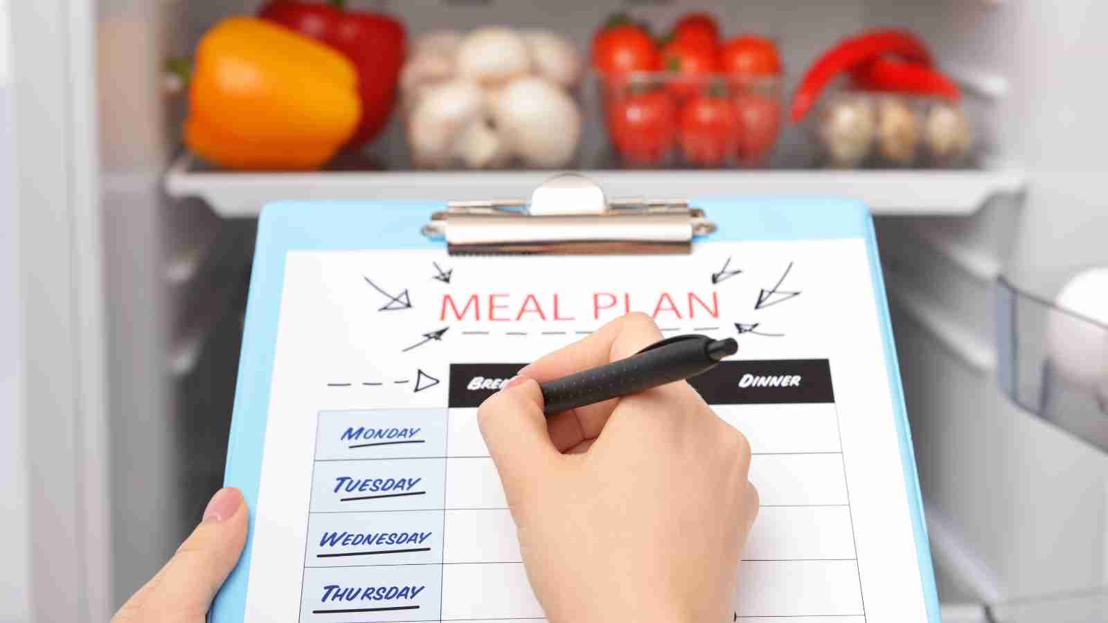 A person writes on a meal plan clipboard in front of an open refrigerator containing vegetables like bell peppers, tomatoes, and mushrooms.