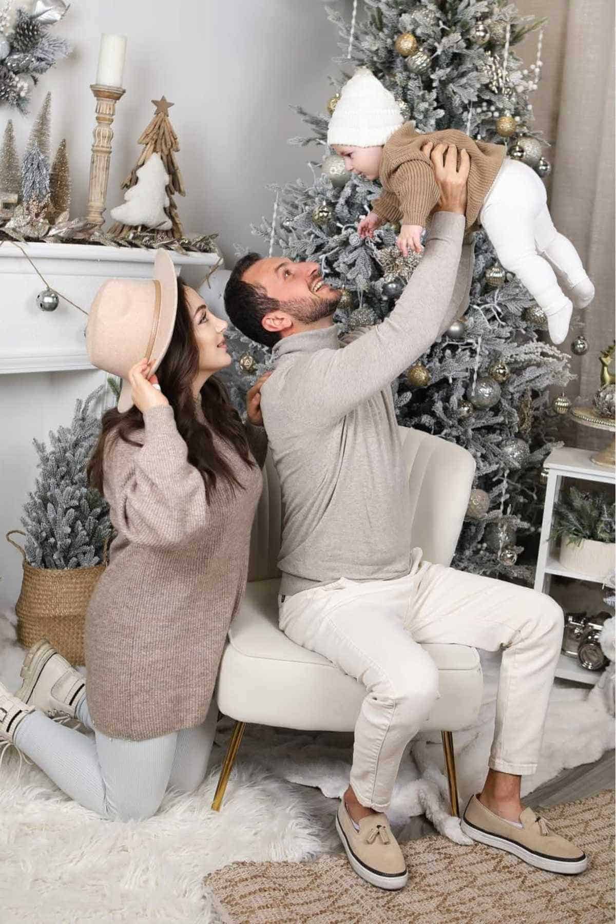 A man and woman pose in front of a decorated Christmas tree as the man lifts a baby dressed in winter clothes. The setting includes festive decor and a cozy atmosphere.