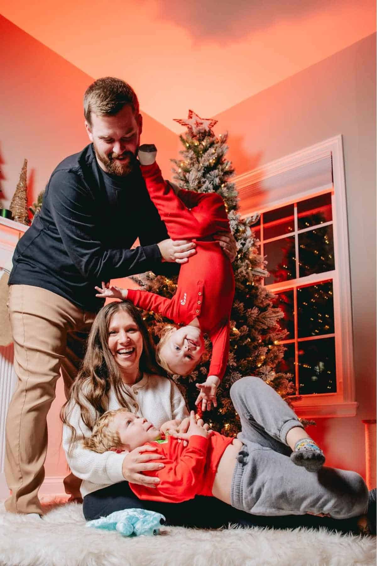 A man playfully holds a child upside down next to a smiling woman and another child lying on the floor, with a Christmas tree and festive decor in the background.
