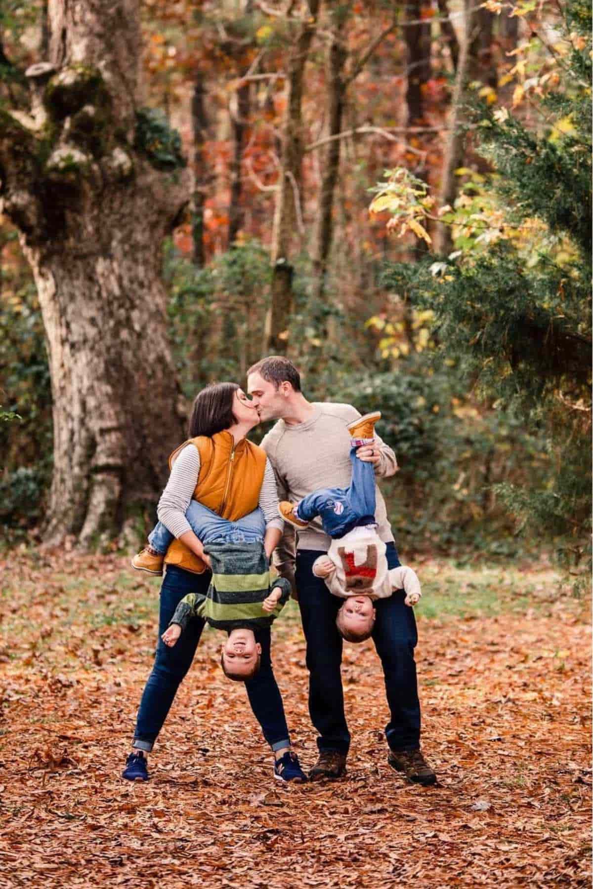 Two adults kiss while each holds a child upside down by the legs in a wooded area with fallen leaves on the ground.
