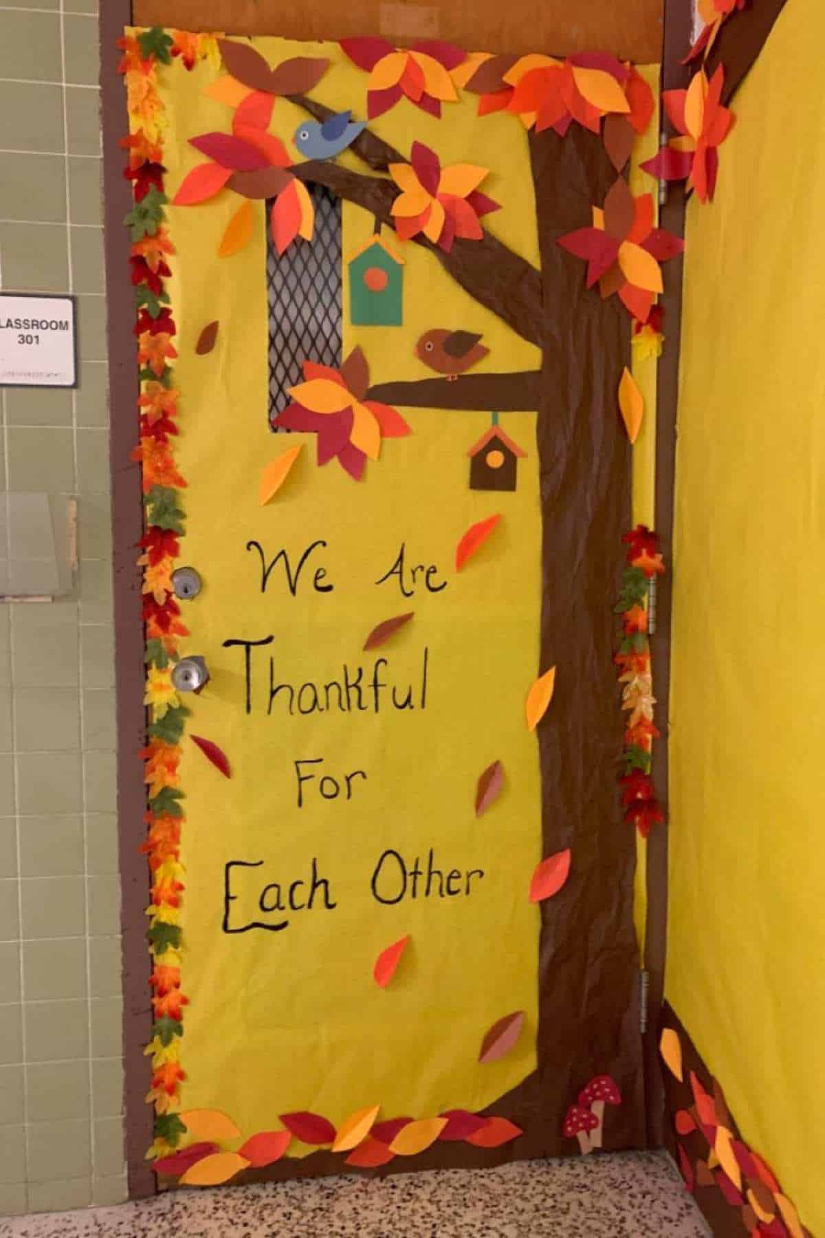 Classroom door decorated with a fall theme, featuring a tree, birds, leaves, and the message "We Are Thankful For Each Other.