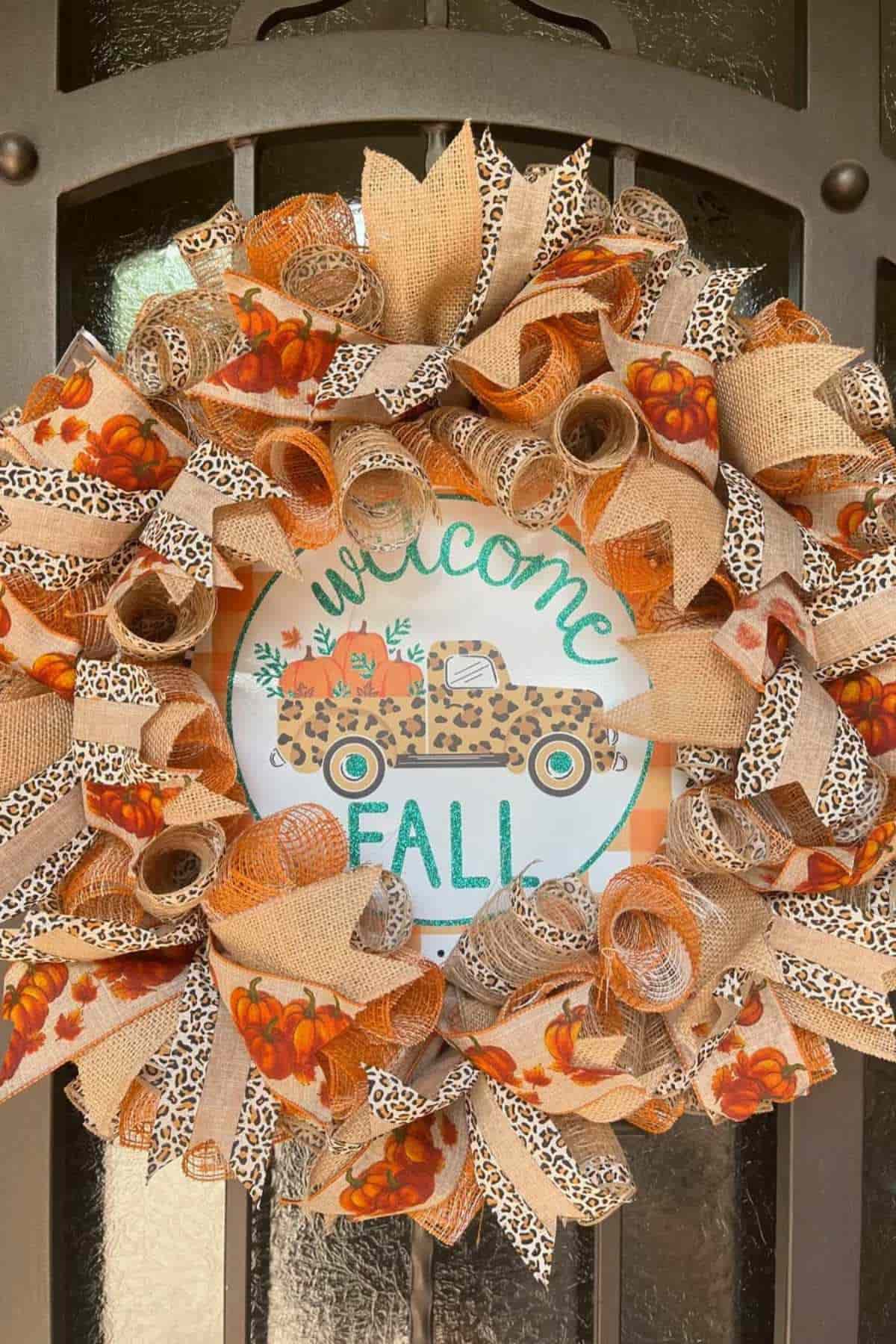 A decorative wreath with orange, beige, and leopard-print ribbons surrounds a sign that reads "Welcome Fall" with a leopard-print truck and pumpkins.