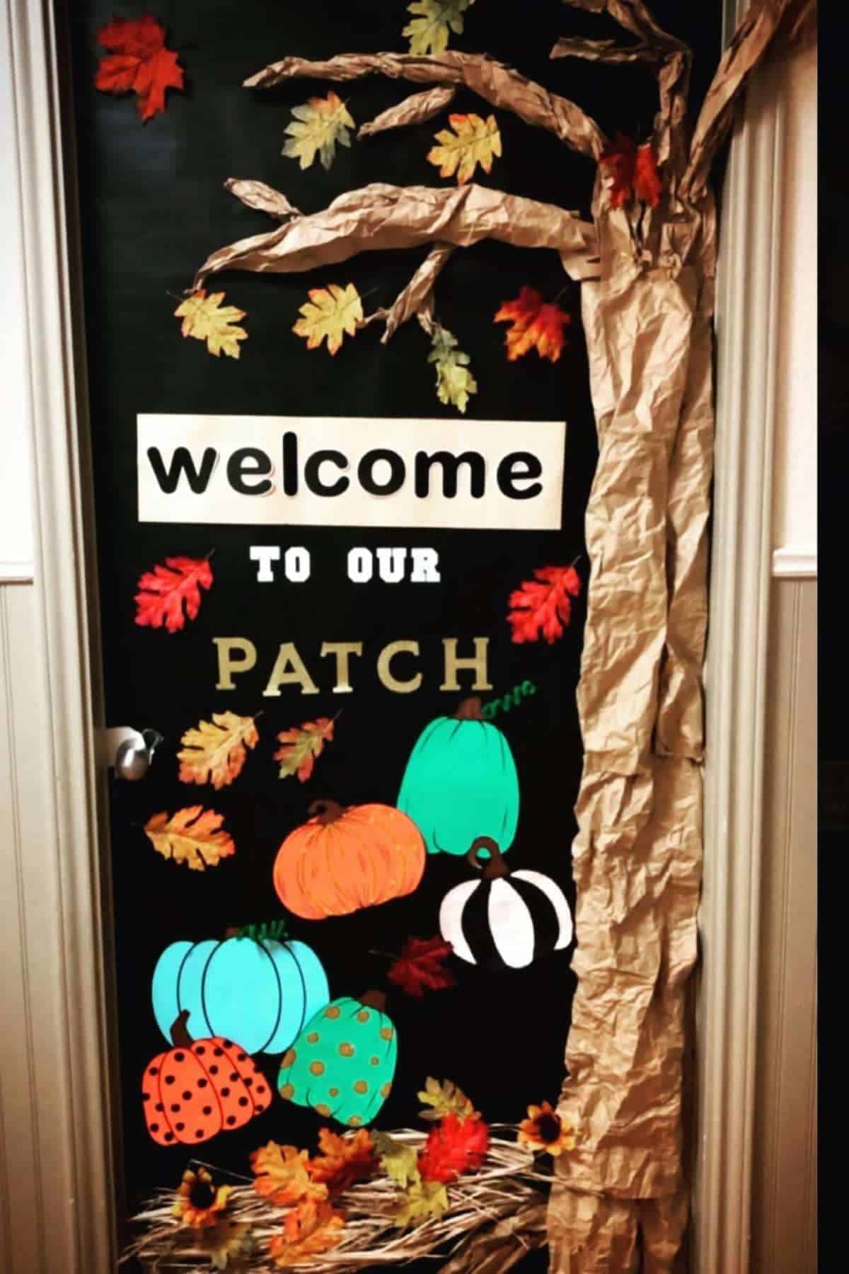 A classroom door decorated with a paper tree, colorful leaves, and pumpkins, with the words "Welcome to our patch" displayed on the door.