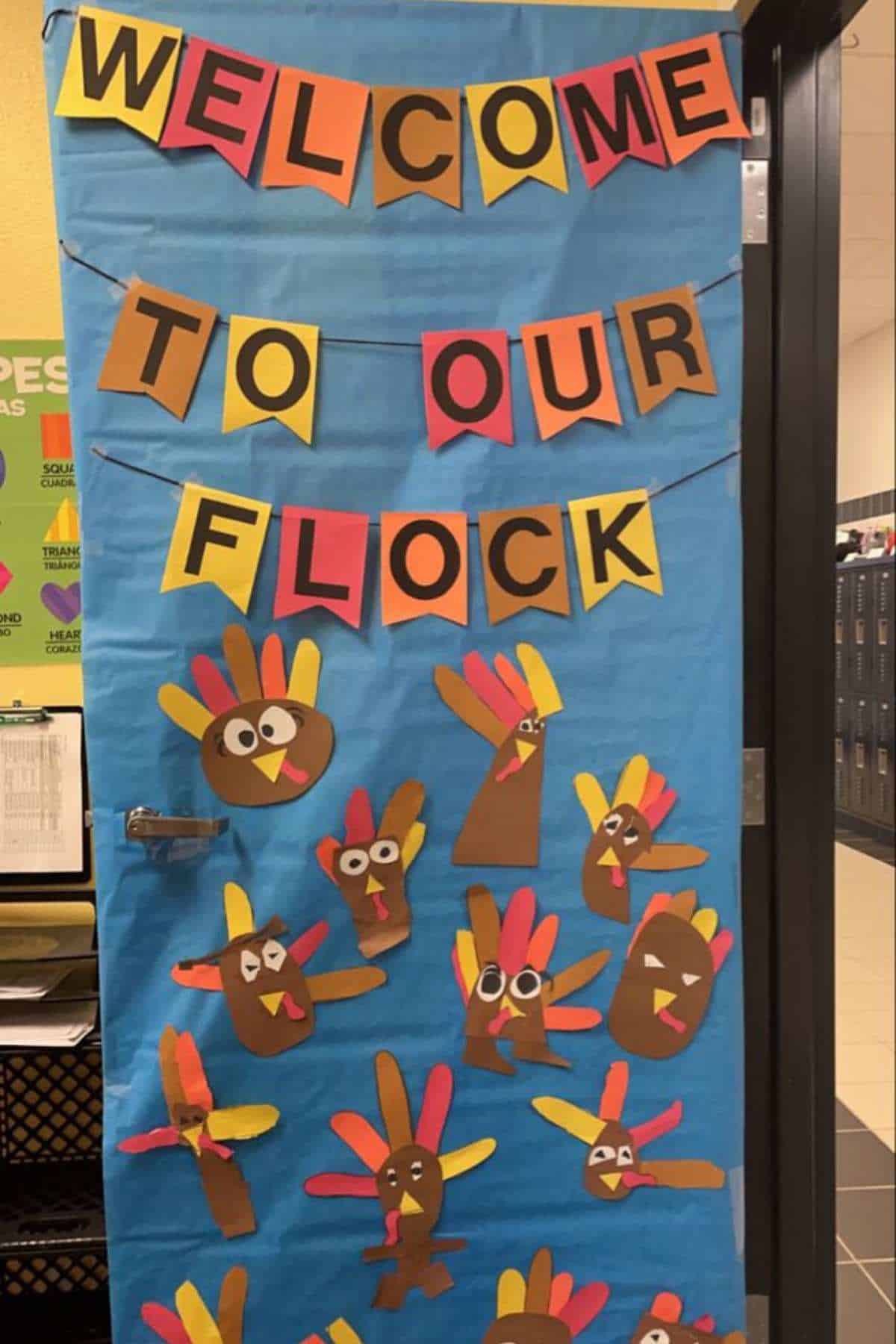 A blue door decorated with paper turkeys made from handprints and a banner reading “Welcome to Our Flock” in colorful letters.