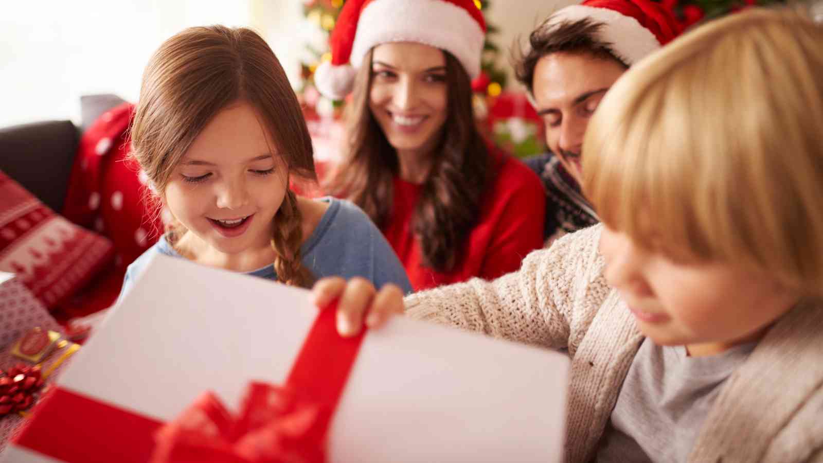 Two children open a gift box with a red ribbon while two adults, both wearing Santa hats, smile in the background.