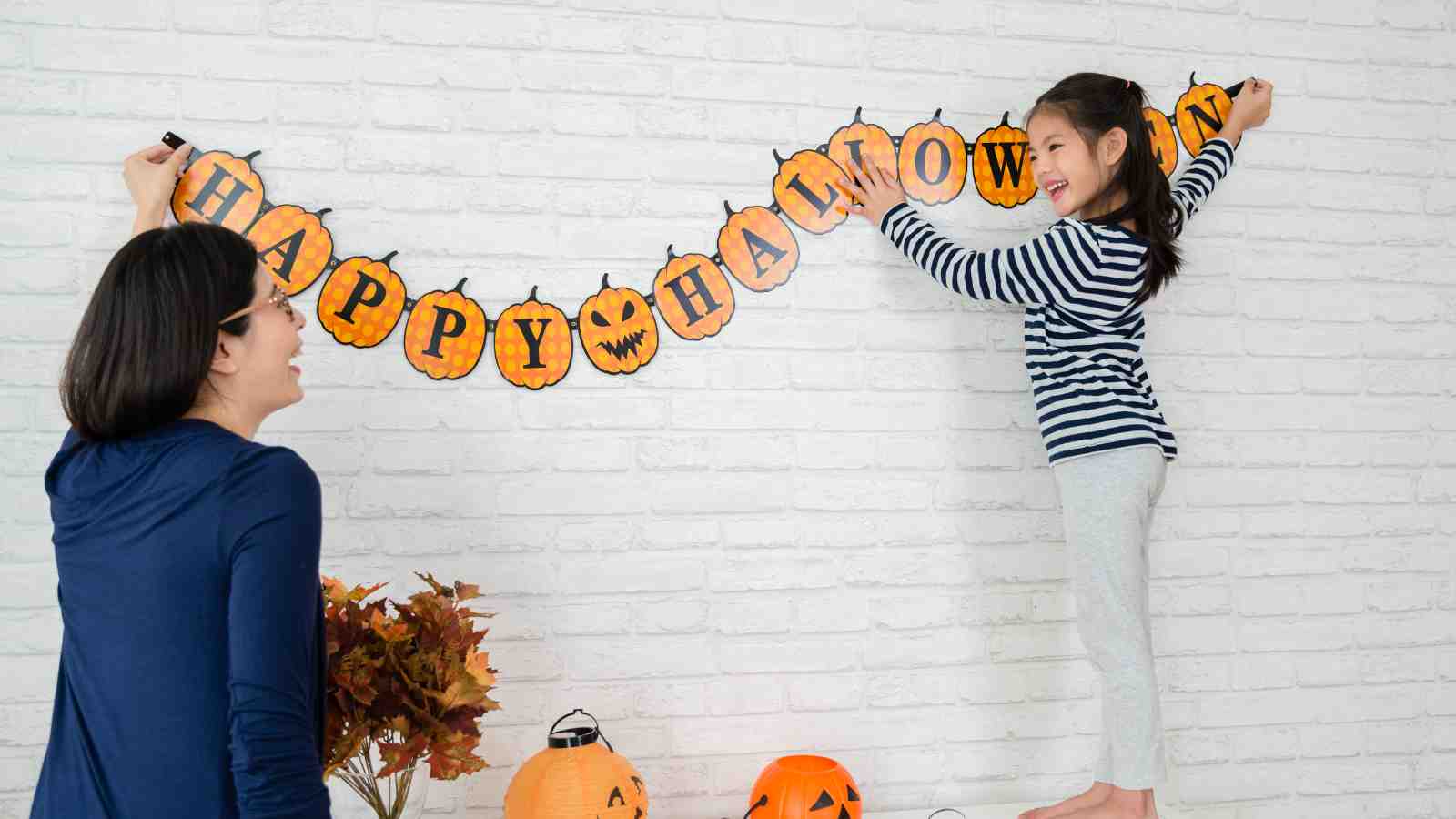 A woman and a girl hang a "Happy Halloween" pumpkin banner on a white brick wall, with jack-o'-lanterns and autumn leaves below.