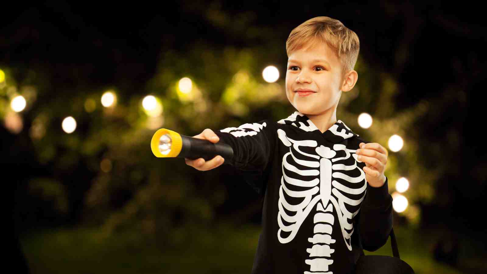 A young boy in a skeleton costume holds a flashlight and smiles outdoors at night, with blurred lights in the background.