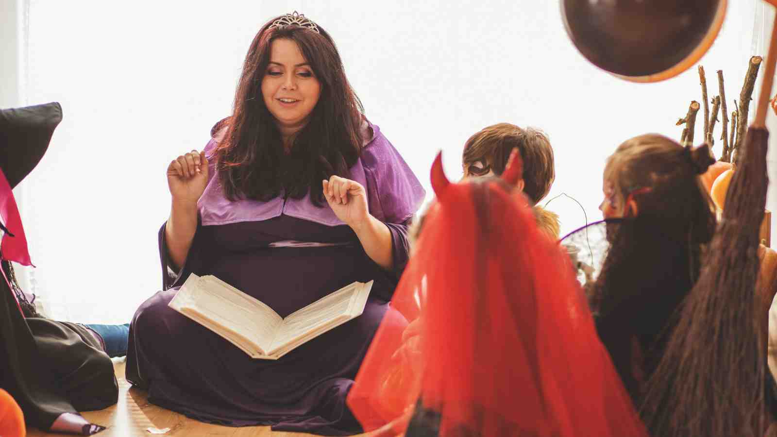 A woman in a purple costume and tiara sits on the floor reading a large book to children dressed in Halloween costumes.