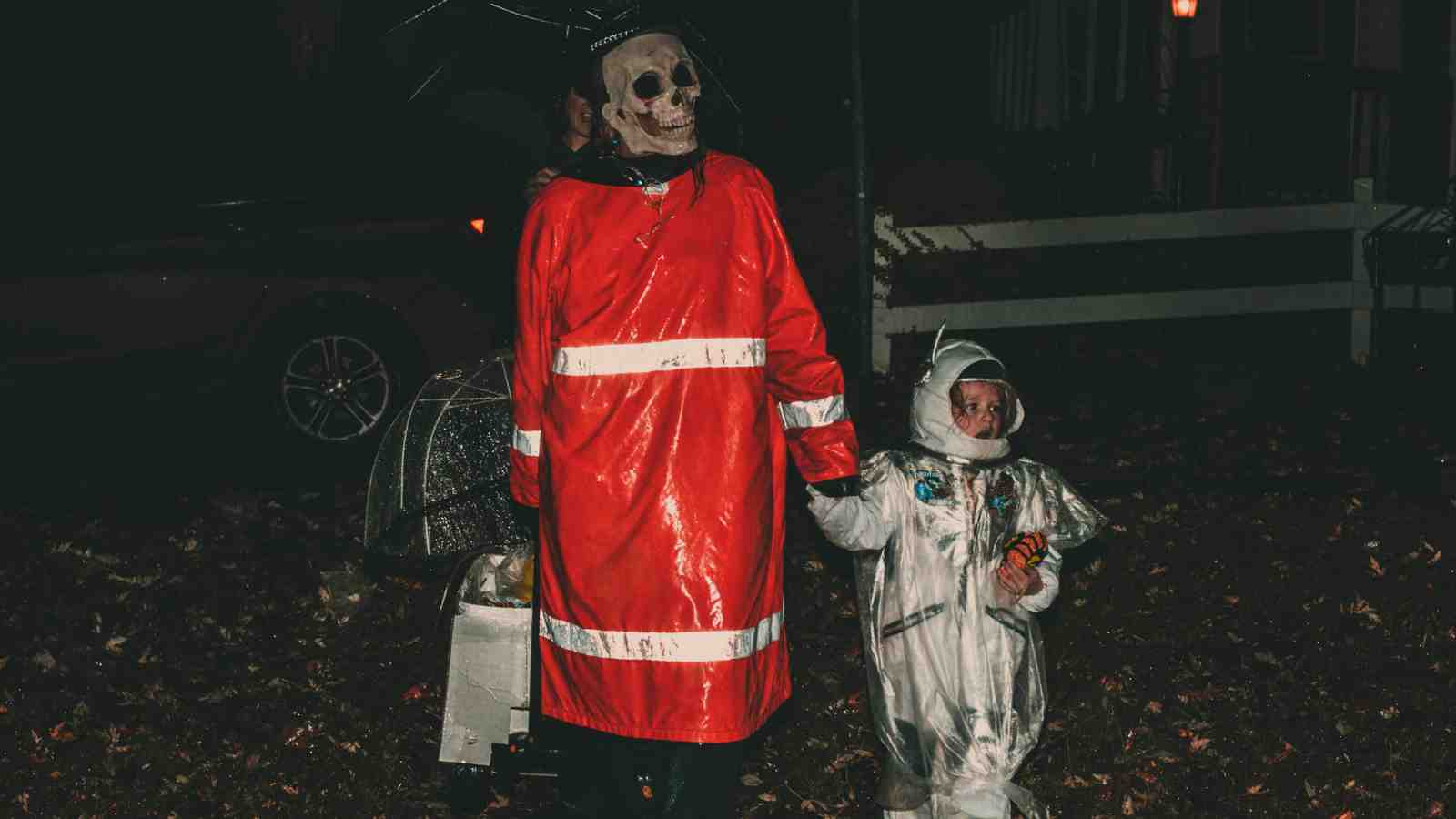 An adult in a skull mask and red raincoat holds hands with a child dressed in a silver astronaut costume, standing outdoors at night.