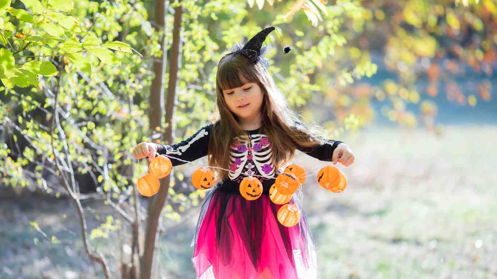 A young girl in a skeleton and pink costume with a witch hat holds a string of small pumpkin lanterns outdoors among trees.