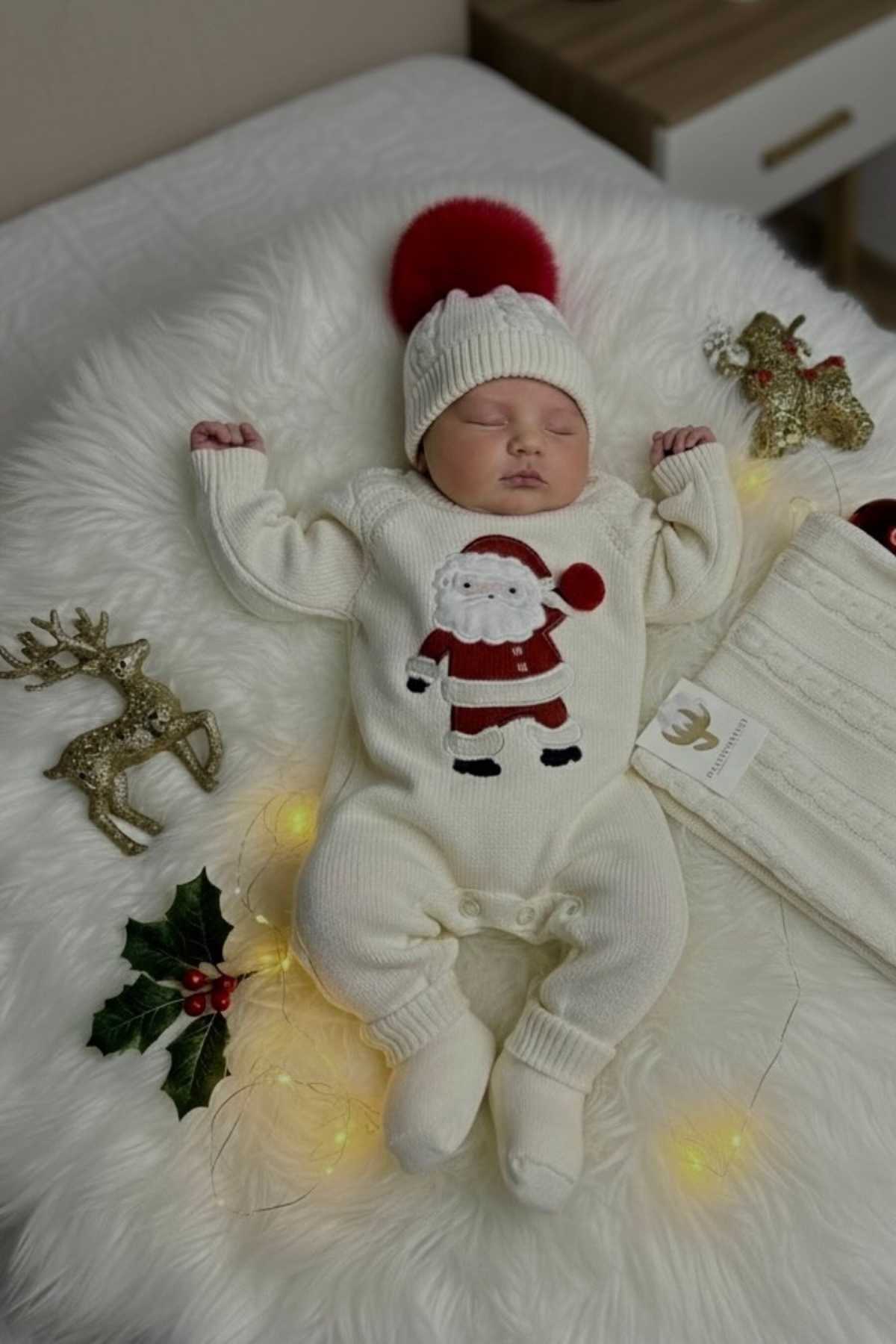 A baby dressed in a white Santa-themed outfit and hat sleeps on a fluffy white blanket, surrounded by holiday decorations and soft yellow lights.