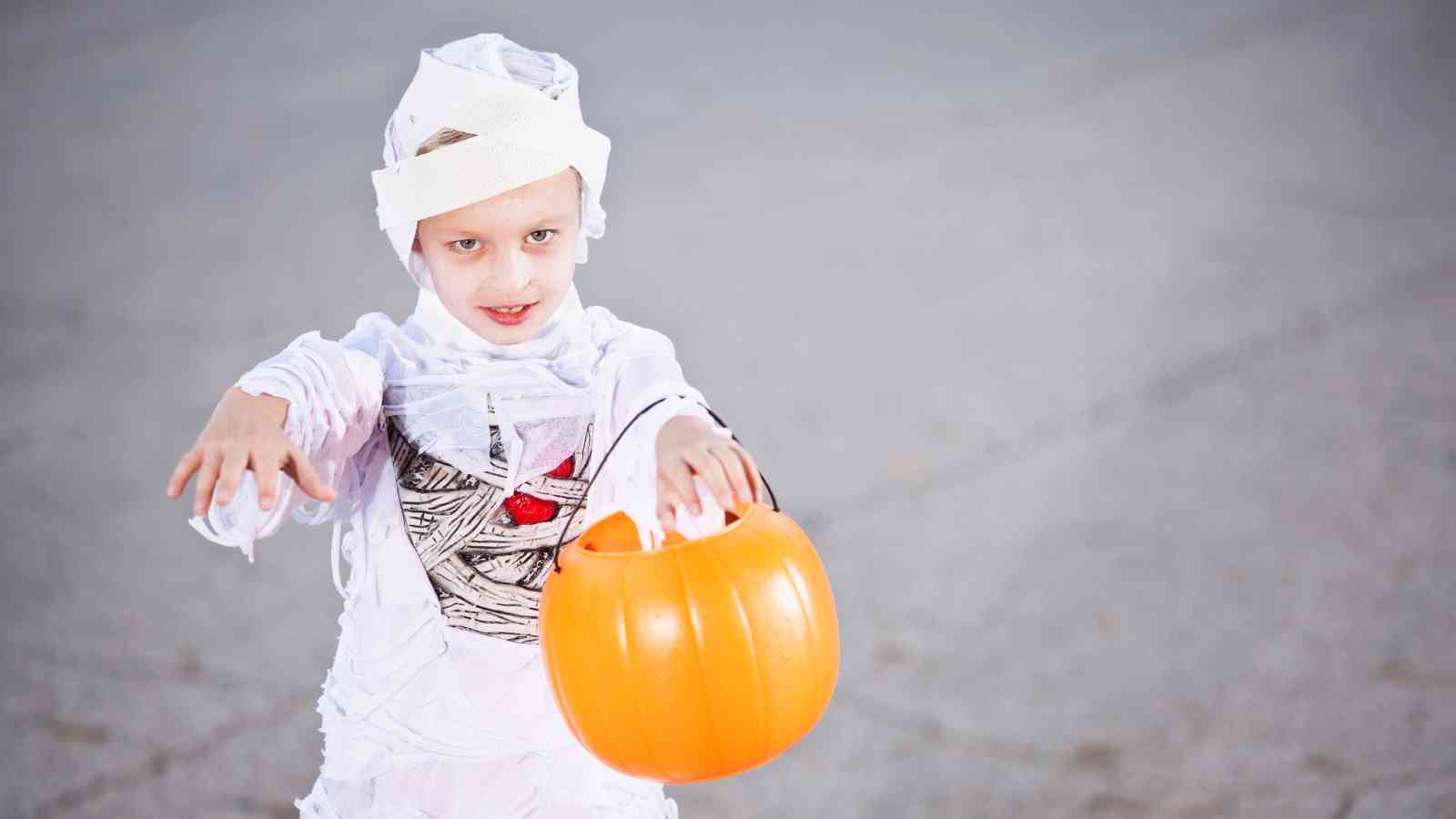 Child in a mummy costume holds an orange pumpkin-shaped bucket and extends their arms forward, standing on a gray surface.