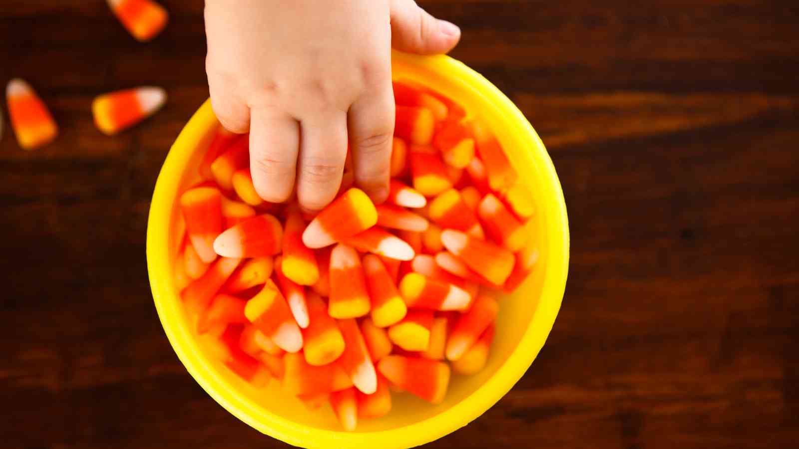 A child's hand reaches into a yellow bowl filled with candy corn on a wooden surface.